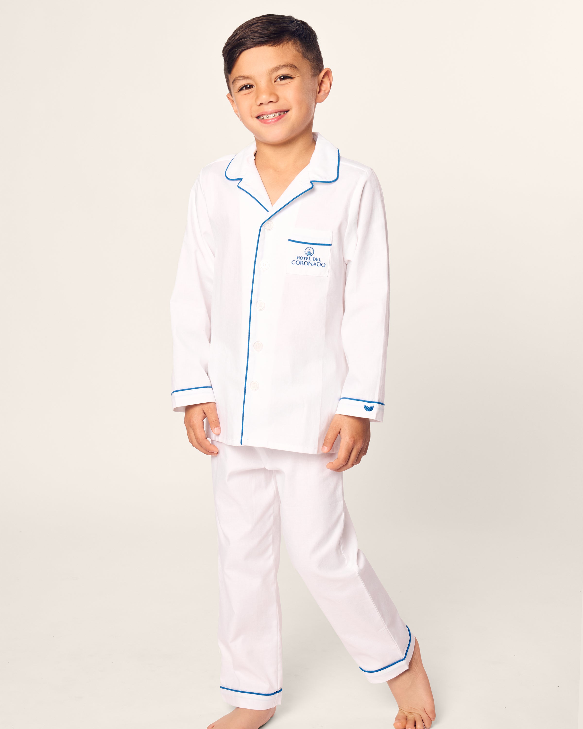 A smiling young boy stands barefoot in the limited-edition Kids Twill Pajama Set Hotel del Coronado x Petite Plume by Petite Plume, featuring elegant white fabric with blue trim, crafted from premium quality cotton against a plain background.