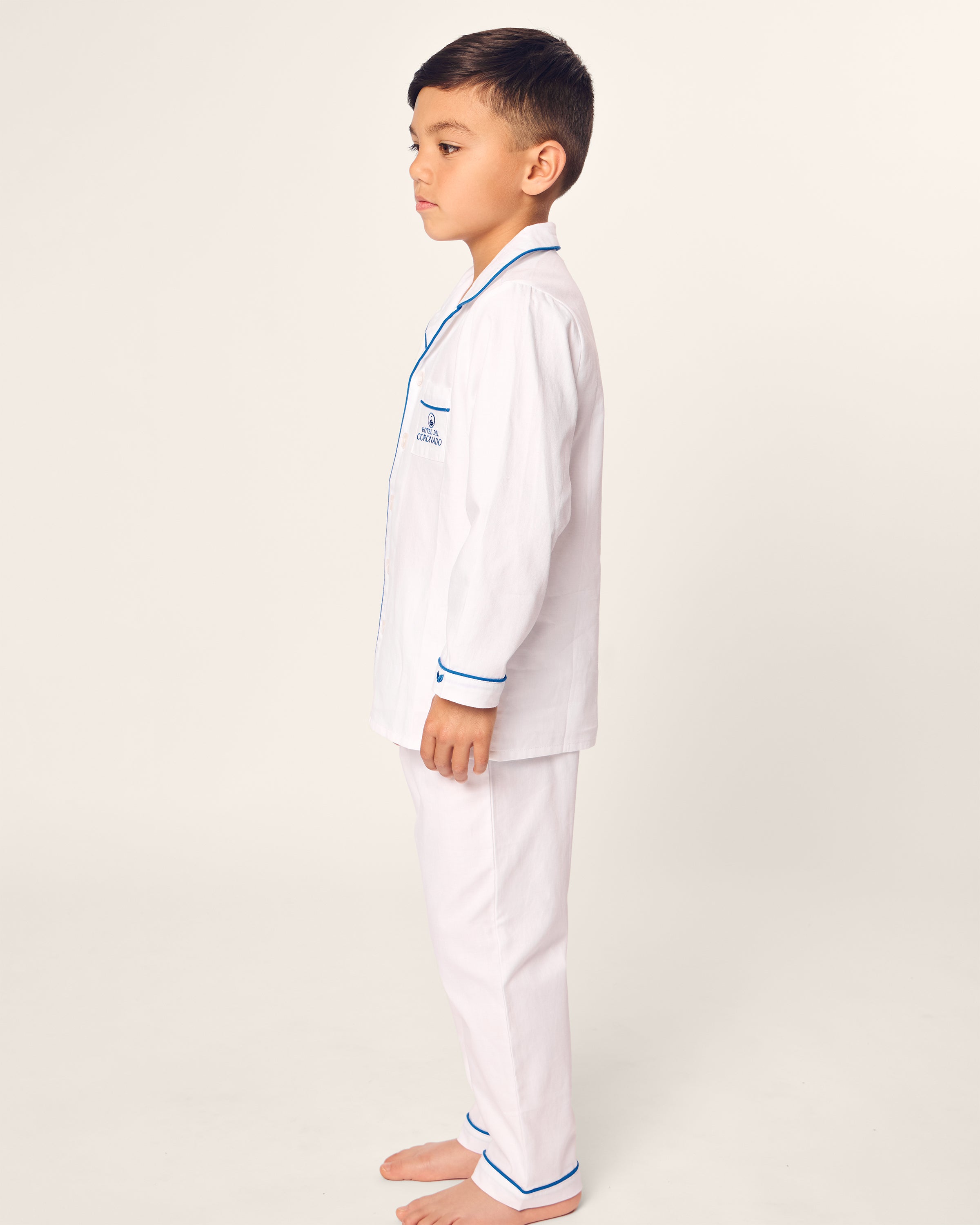 A young boy, barefoot in a side profile, wears a Kids Twill Pajama Set Hotel del Coronado x Petite Plume by Petite Plume. The elegant set features blue trim and an embroidered plane on the chest, made from the finest quality cotton for ultimate comfort against a plain background.