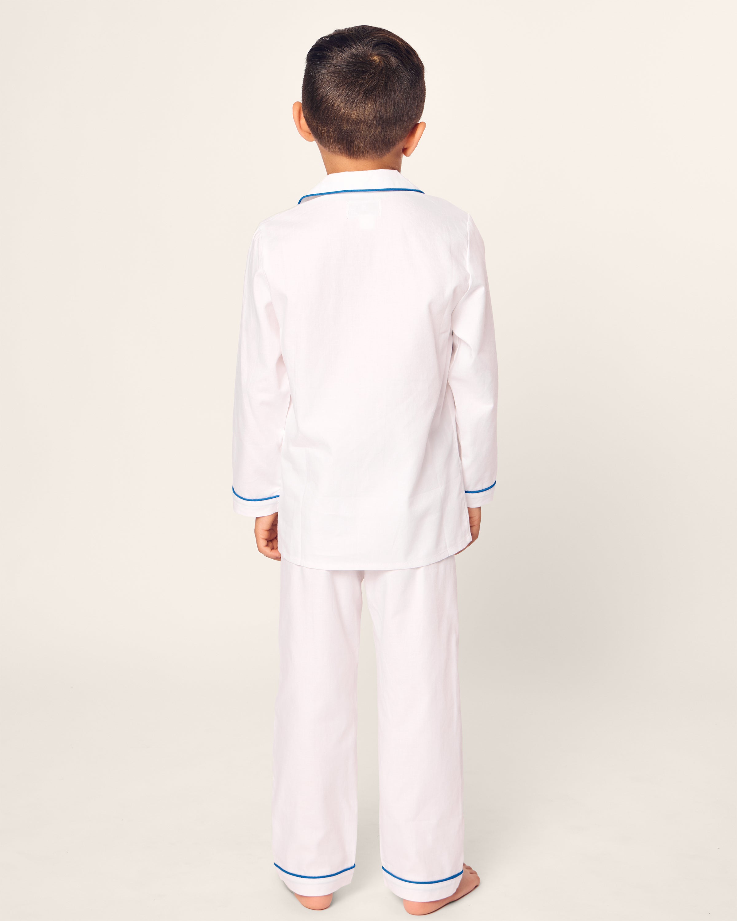 A child is seen from the back wearing the Kids Twill Pajama Set Hotel del Coronado x Petite Plume in light pink with blue piping, crafted from premium cotton by Petite Plume. The limited-edition set includes long sleeves and pants, perfect for stylish sleepwear.