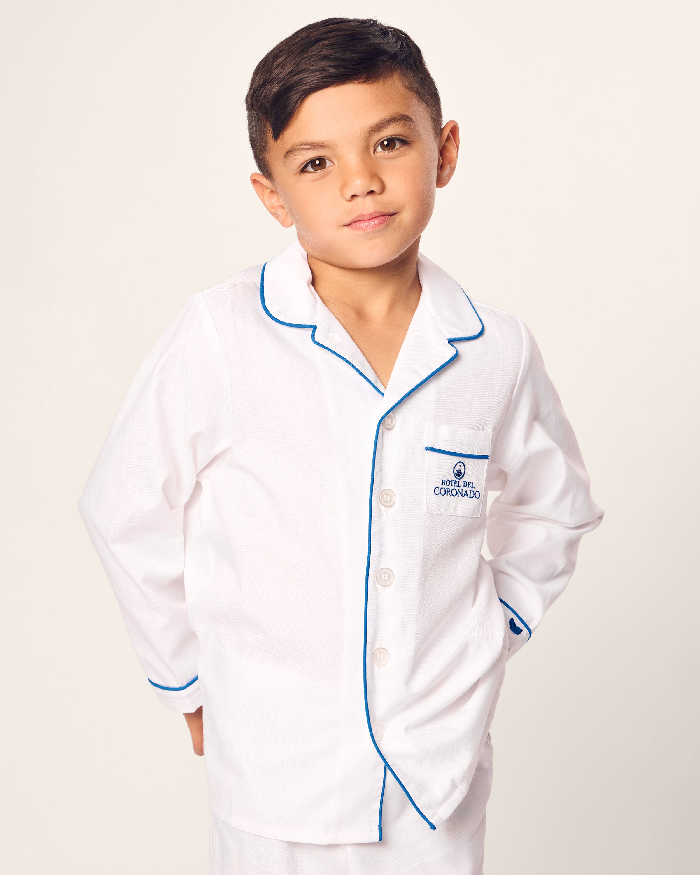 A boy confidently poses in a Hotel del Coronado x Petite Plume Kids Twill Pajama Set by Petite Plume. The elegant white pajamas, made of the finest cotton with blue piping and a chest pocket logo, exude charm. With short dark hair and hands behind his back, he stands against a plain backdrop.