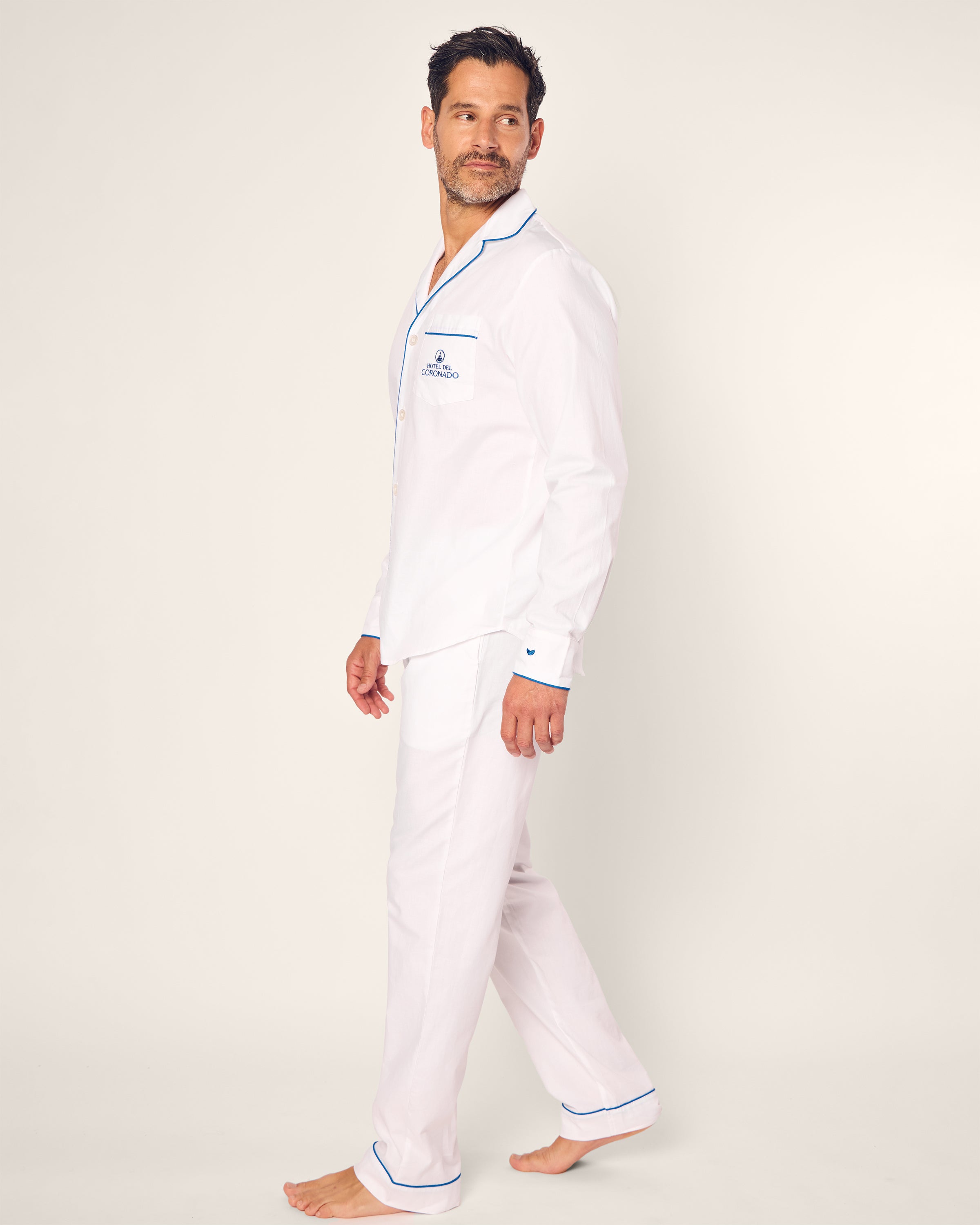 A man stands barefoot, gazing off to the side in a Mens Twill Pajama Set Hotel del Coronado x Petite Plume, featuring blue trim and an embroidered chest pocket logo. This collaboration exudes elegance against a plain, light-colored backdrop.