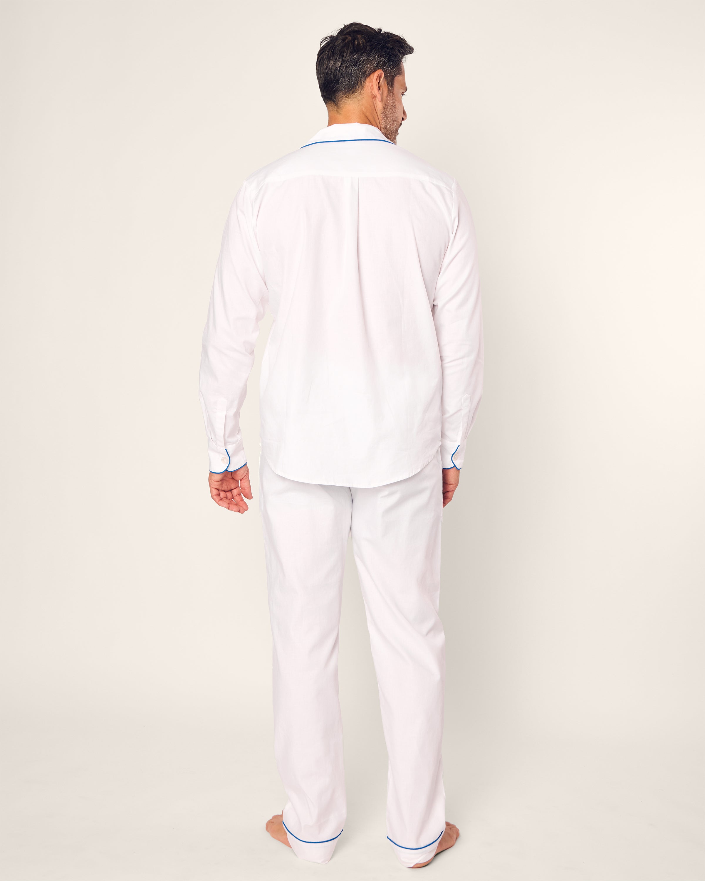 A man, barefoot against a plain white background, wears the luxurious Mens Twill Pajama Set from the Hotel del Coronado x Petite Plume collaboration.