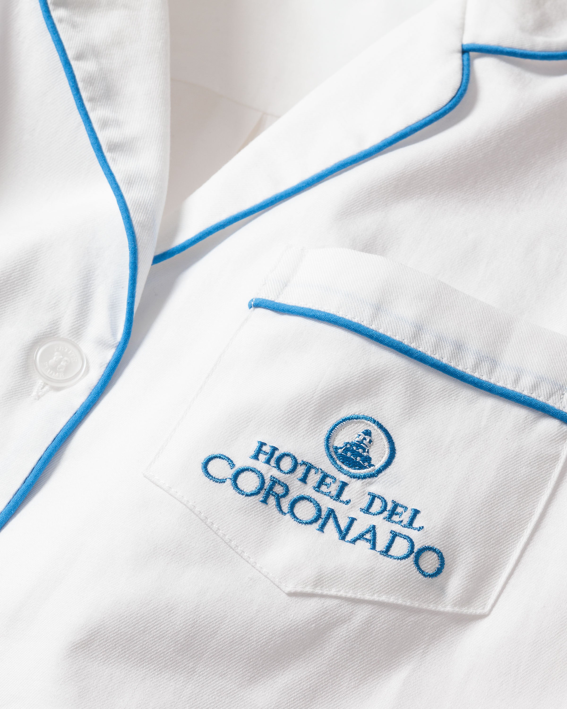 This elegant Petite Plume x Hotel del Coronado pajama set for men features white fabric with blue piping and embroidery. The visible pocket reads HOTEL DEL CORONADO, with a circular emblem above, while a white button adorns the left piping.