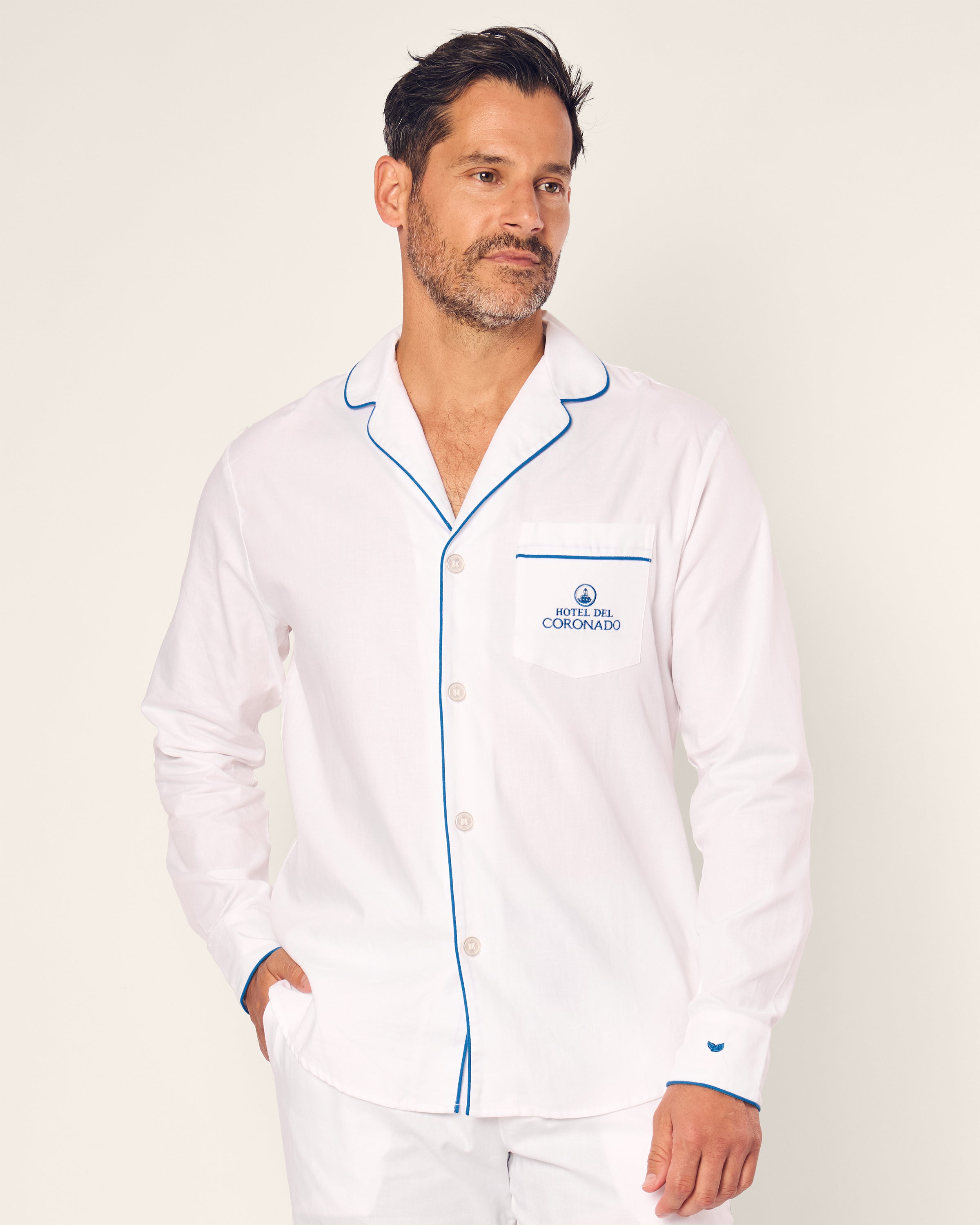 A man stands against a plain background wearing the Petite Plume Mens Twill Pajama Set in white with blue piping, embroidered with a Hotel Colorado logo on the chest. Part of the Hotel del Coronado x Petite Plume collection, he glances to the side with one hand in his pocket.
