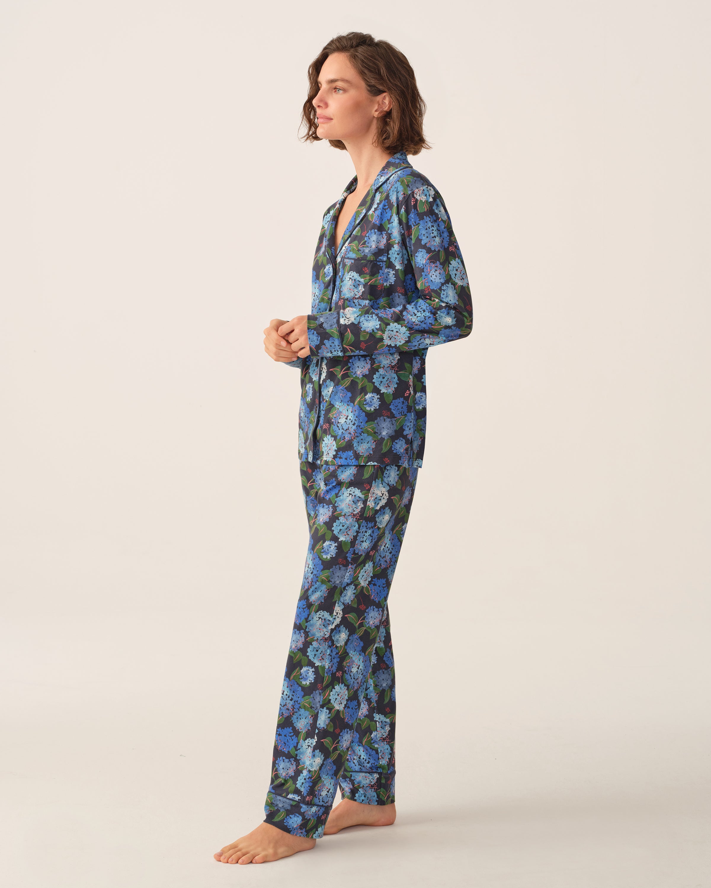 A person stands barefoot in profile against a plain, light backdrop, wearing the Petite Plume x Hedges Women’s Pima Pajama Set in Hydrangea—a long-sleeved, button-up pajama set featuring a blue and purple floral pattern on a black background.