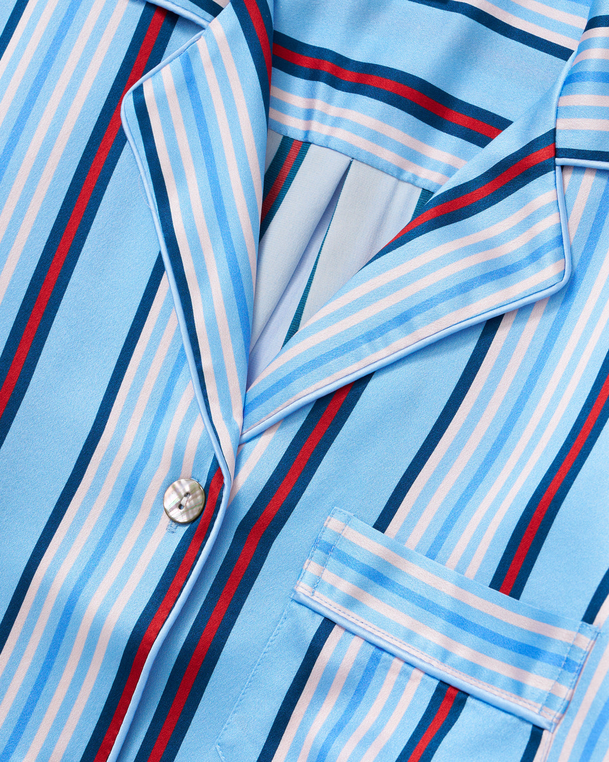 Close-up of the Petite Plume x Hedges Women’s Silk Short Set in Blue Stripe, featuring a button-up pajama shirt with blue, white, red, and light pink stripes, a front pocket, a collar, and crafted from silky fabric.