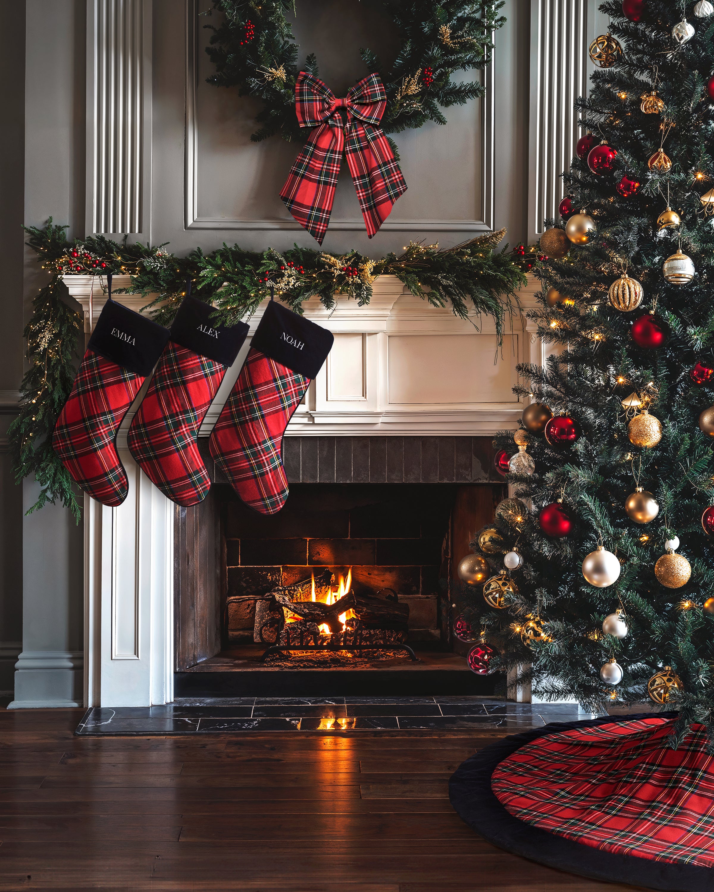 A cozy living room has a lit fireplace with Petite Plumes monogrammable cotton Christmas Stocking in Imperial Tartan. A red-and-gold dressed tree adds charm, while a Petite Plume holiday print plaid wreath with a bow above the mantel seals the festive atmosphere.