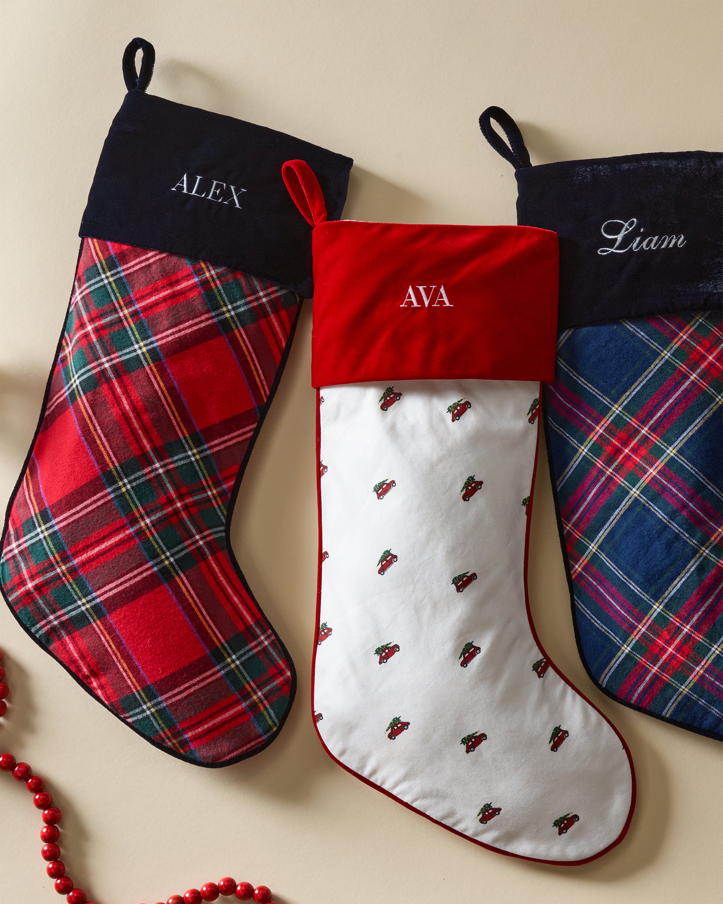 Three holiday stockings hang in a row: the first and third are monogrammable cotton with embroidered names, while the middle is a Petite Plume Imperial Tartan stocking featuring a red top and small holly patterns. Red beads hang below.