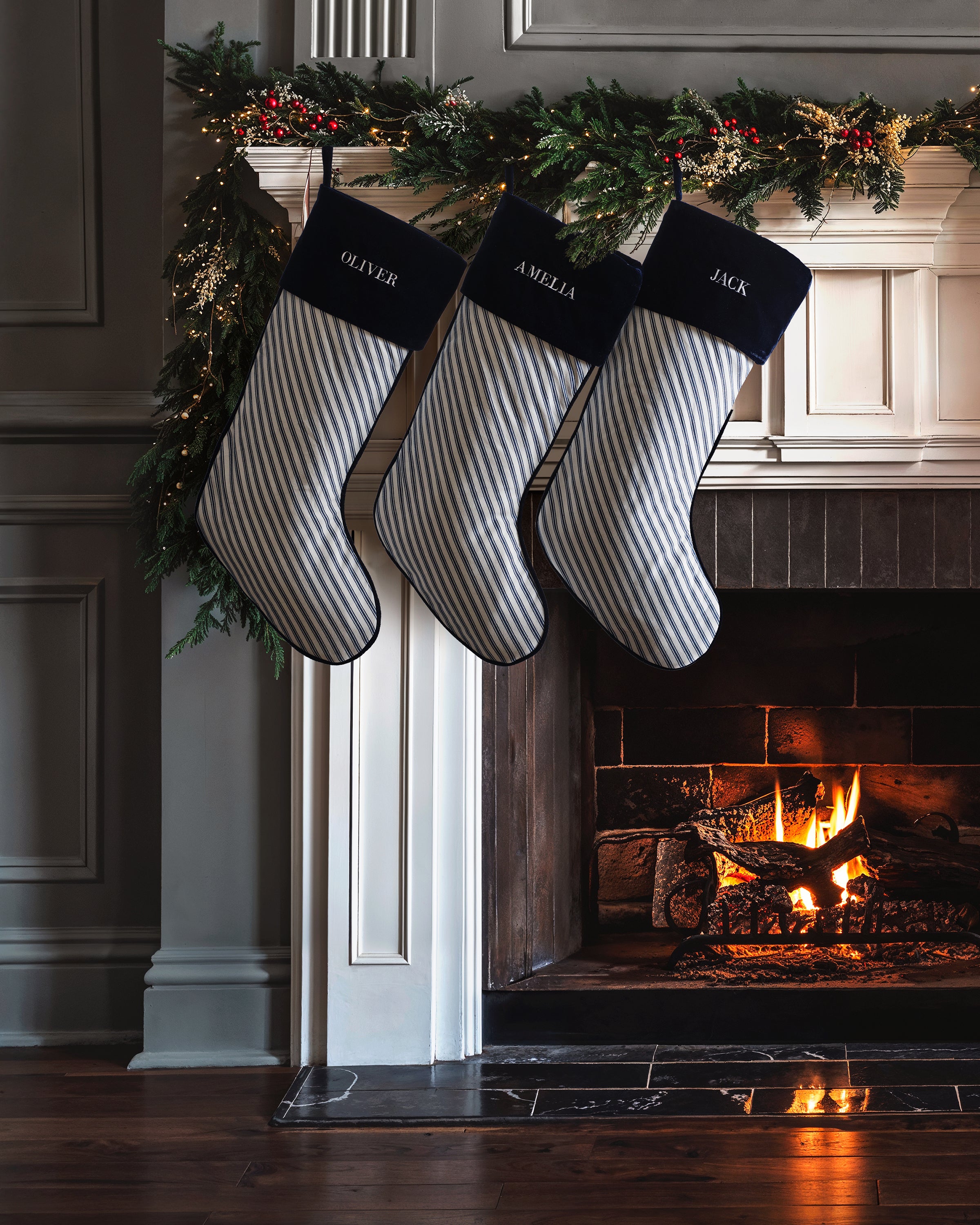Three monogrammable Petite Plume stockings in Navy French Ticking, labeled Oliver, Amelia, and Jack, hang from a mantel adorned with greenery and lights. A cozy fire glows below, enhancing the holiday ambiance.