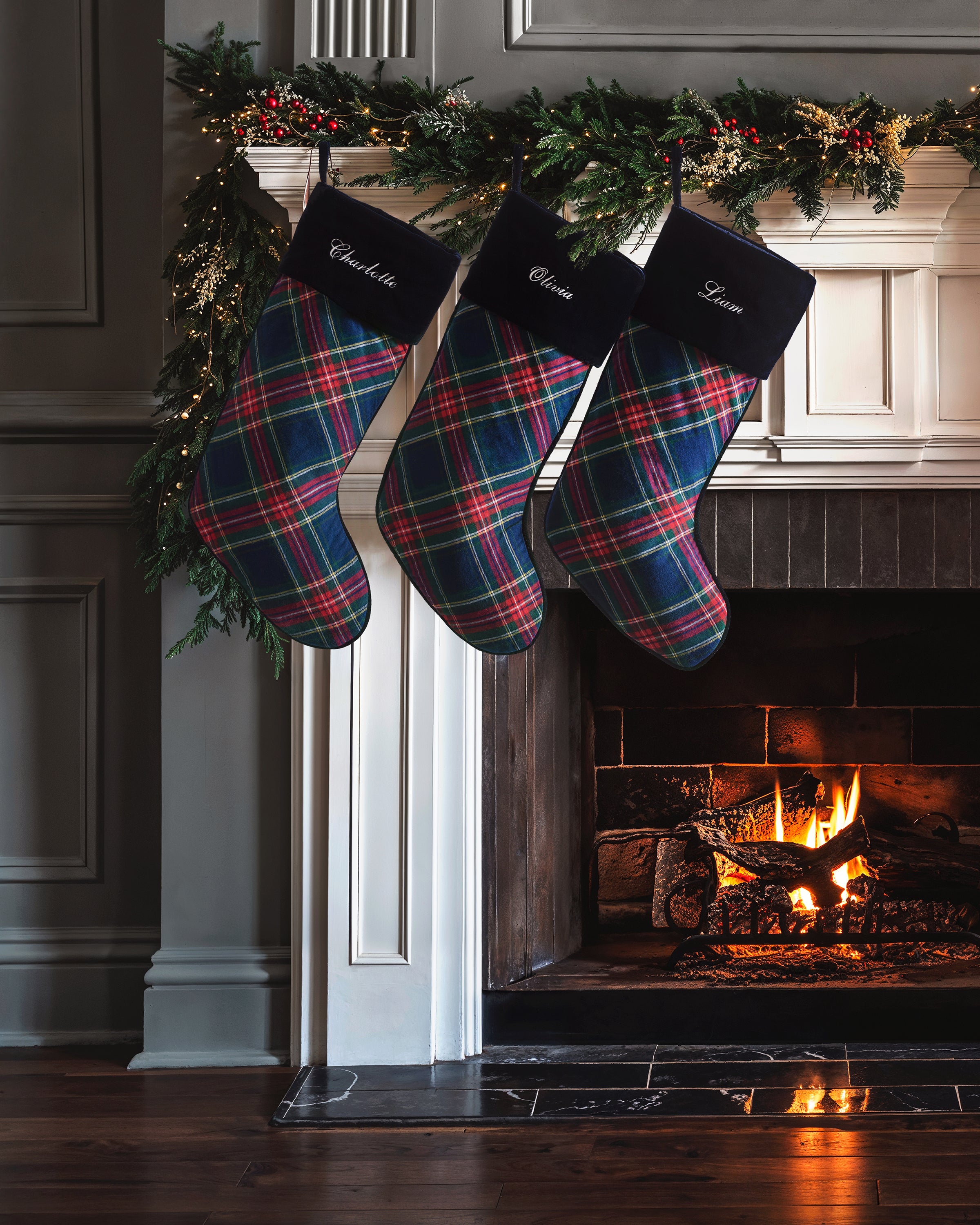 Three Windsor Tartan Christmas stockings by Petite Plume hang from a mantel adorned with greenery and lights. Below, a warm fire crackles, creating a cozy holiday vibe.