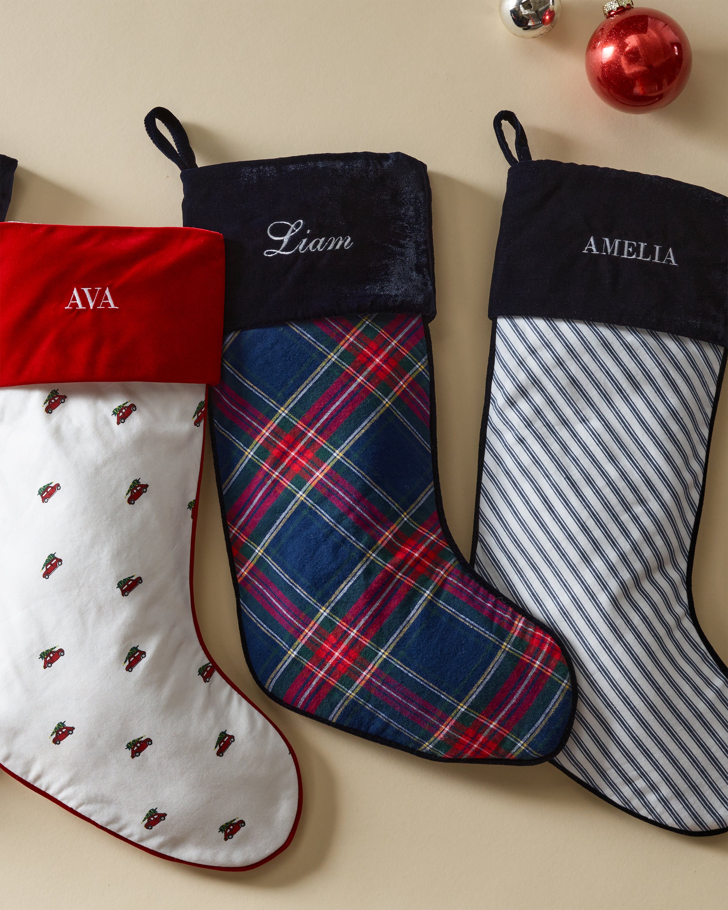 Three personalized holiday stockings hang on the wall: a white and red holly pattern for Ava, Petite Plumes Windsor Tartan for Liam, and a blue striped design for Amelia. A red ornament rests nearby.