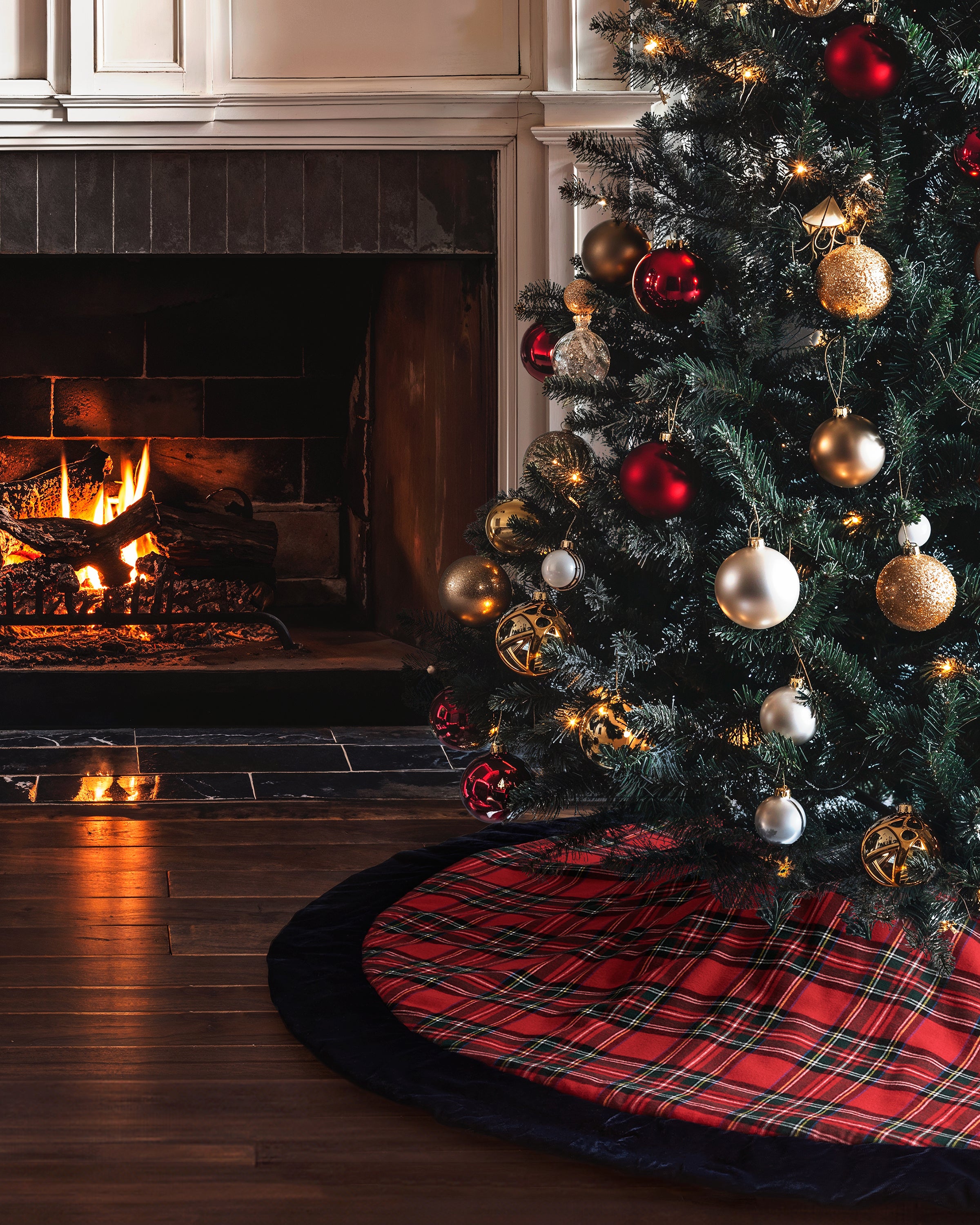 A decorated Christmas tree with red and gold ornaments stands by a lit fireplace on a festive, monogrammed red plaid Christmas Tree Skirt in Imperial Tartan by Petite Plume. The warm fire glow enhances the holiday decor on the polished wooden floor.