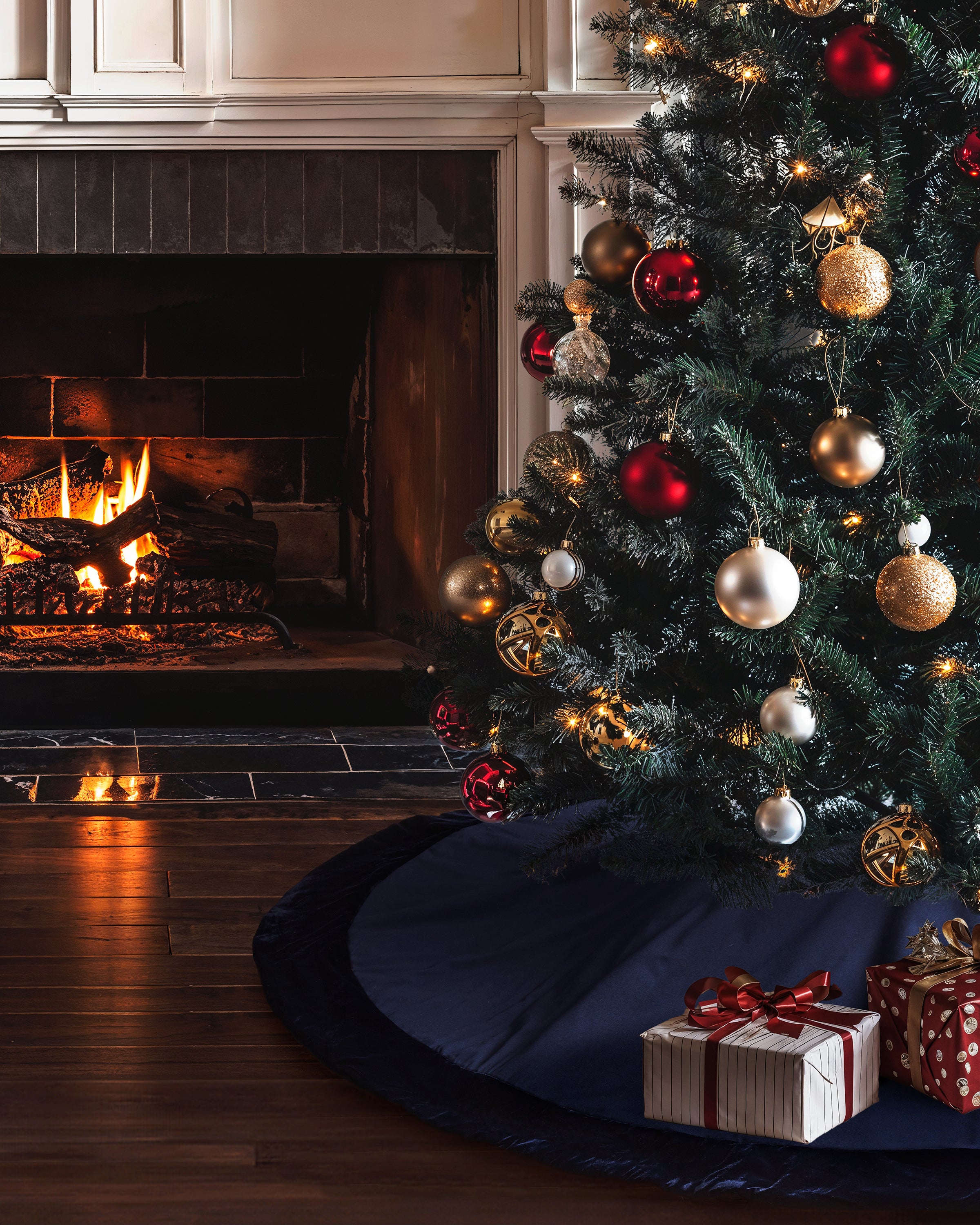 A Christmas tree with a red and gold theme stands proudly on a base elegantly adorned by Petite Plumes navy tree skirt. Wrapped gifts enhance the charm, while a fireplace crackles on the left, spreading a warm glow over the festive decor.