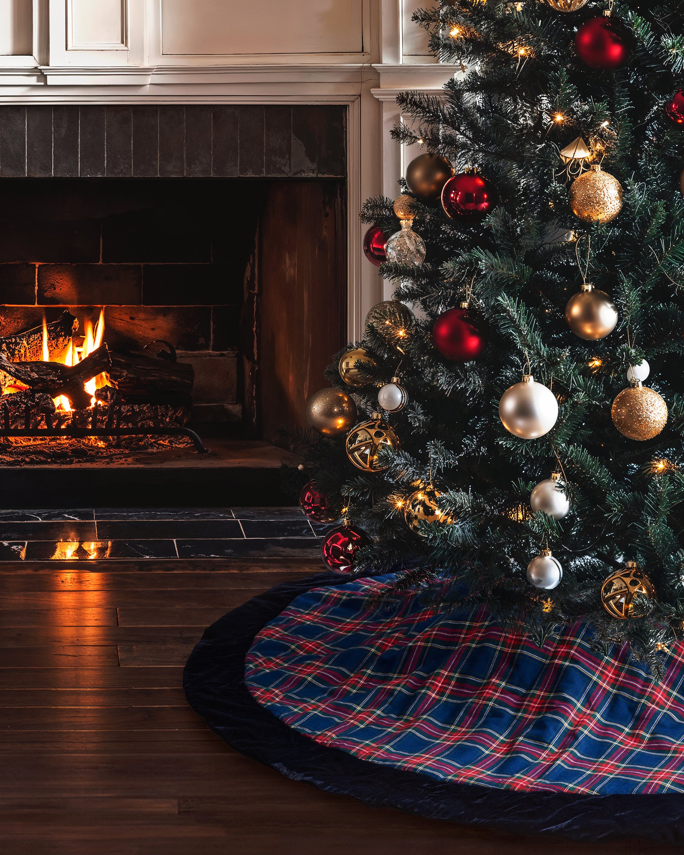A Christmas tree with gold and red ornaments stands by a crackling fireplace. The Petite Plume Christmas Tree Skirt in Windsor Tartan, adorned with glowing lights and intricate holiday decor, adds elegance. Wooden floors enhance the rooms warm, cozy atmosphere.