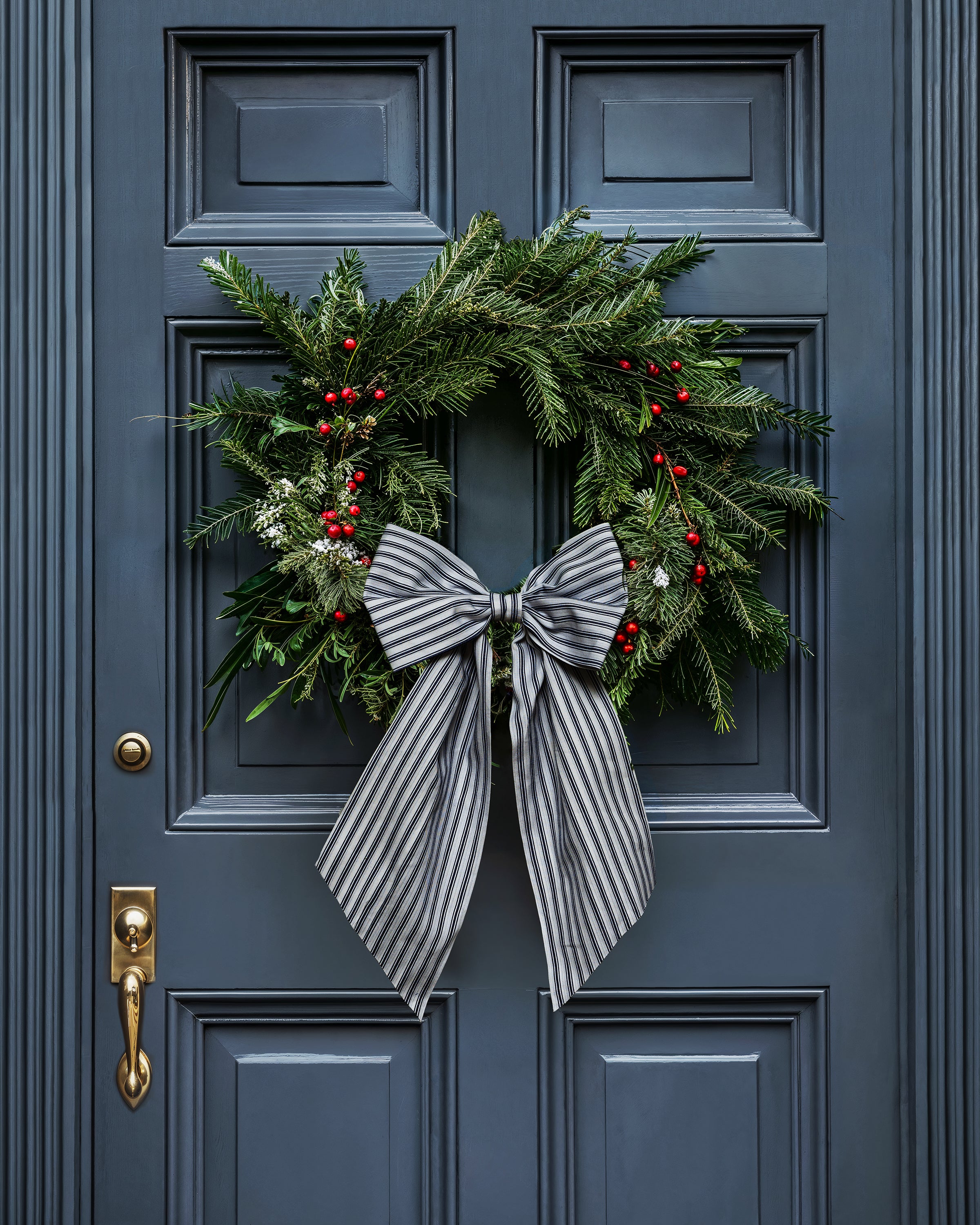 A festive Christmas wreath with green foliage, red berries, and white accents adorns a dark blue door, topped by a striking Petite Plume Wreath Bow in Navy French Ticking. A gleaming gold door handle on the left completes this holiday decor scene.