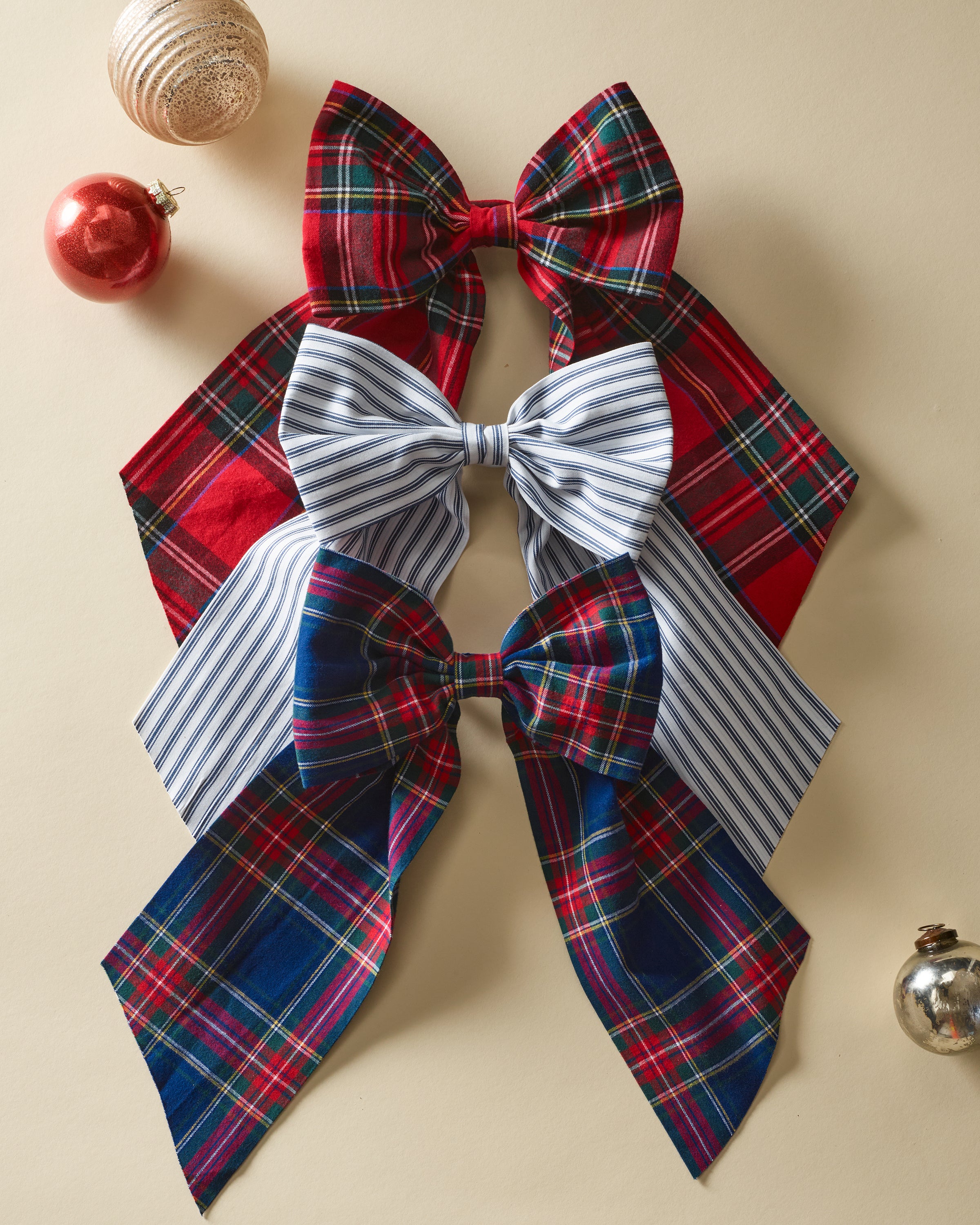 A trio of festive bows from Petite Plume, including a plaid red, a blue and white striped Wreath Bow in Navy French Ticking, and a blue plaid bow, are arranged vertically on beige. Holiday ornaments scattered around them add cheer to any setting.