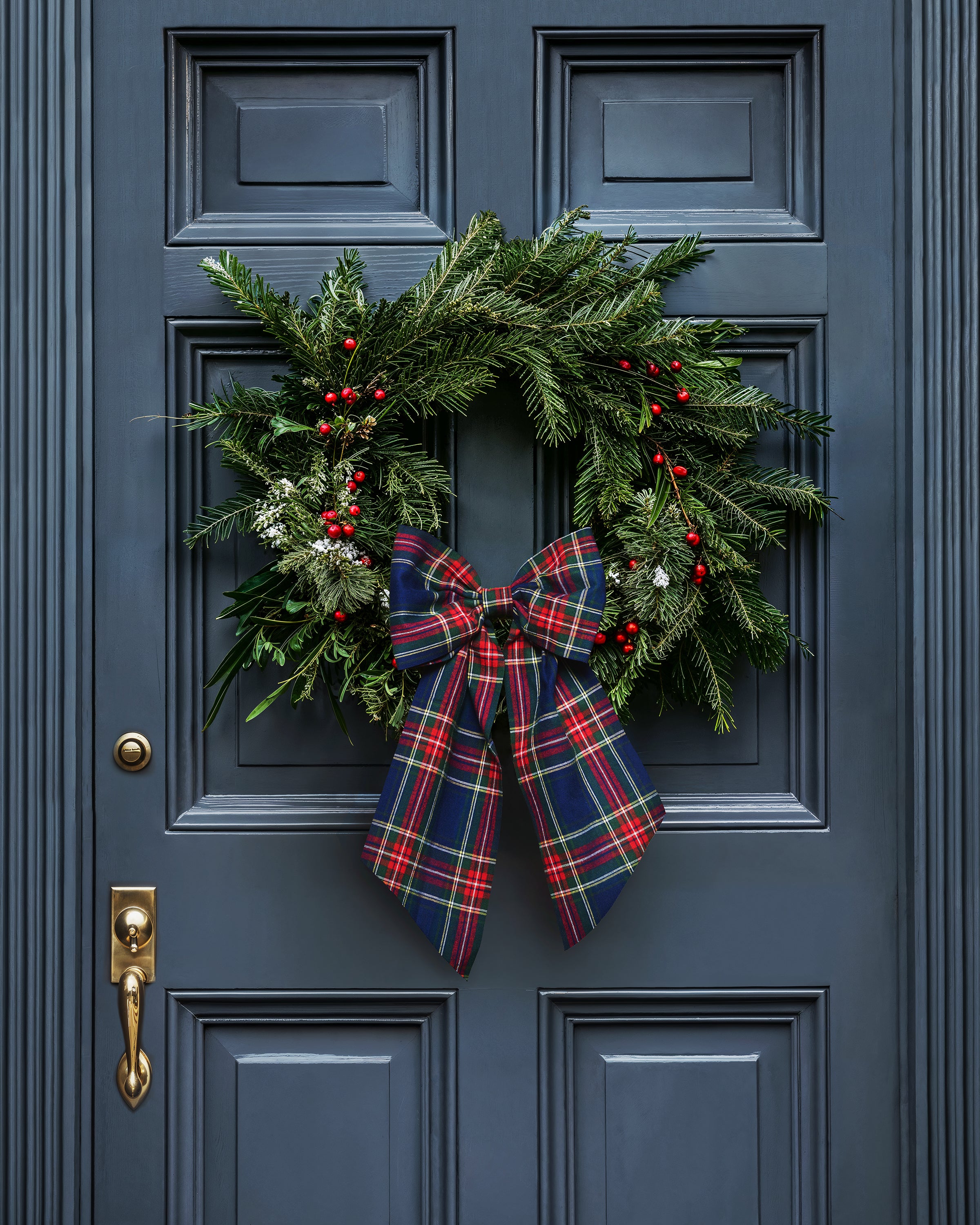 A festive wreath with evergreen branches and red berries, adorned with Petite Plumes Wreath Bow in Windsor Tartan, hangs on the dark blue door as perfect holiday decor. Below it, a brass door handle gleams subtly in the background.