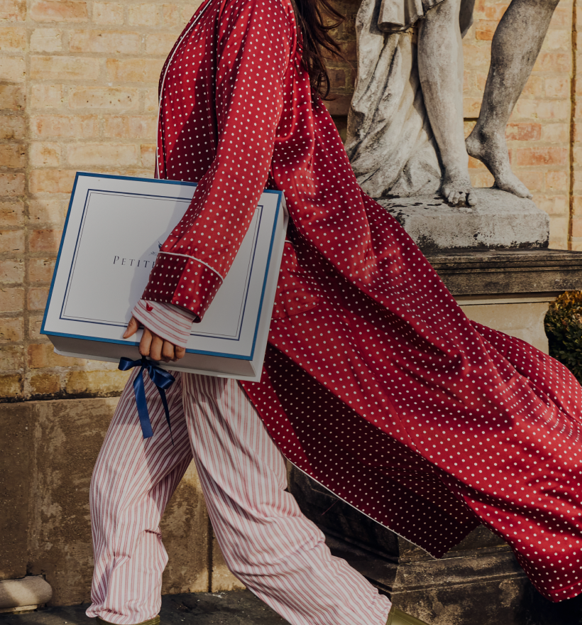 A person in red polka-dot pajamas and a matching robe walks outside, holding a large gift box with a blue ribbon. Only the lower half of their face is visible. A stone statue and brick wall are in the background. A person in red polka-dot pajamas and a matching robe walks outside, holding a large gift box with a blue ribbon. Only the lower half of their face is visible. A stone statue and brick wall are in the background.