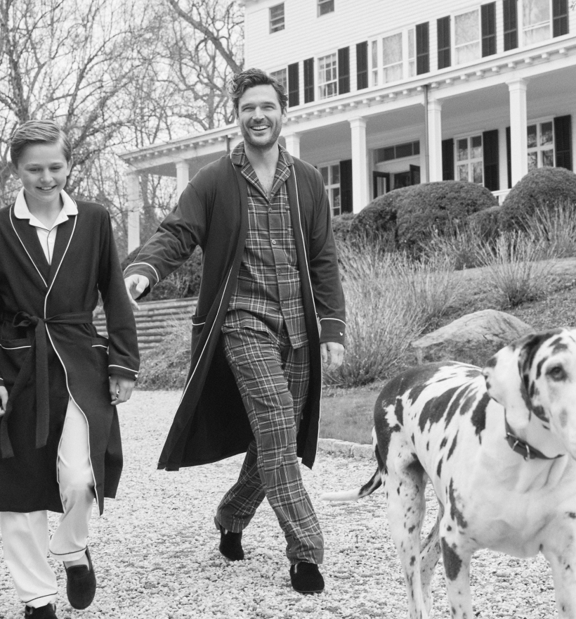 A black-and-white photo of two people wearing pajamas and robes walking outside near a large house, smiling, with a Great Dane dog walking in the foreground. Leafless trees and bushes are visible in the background. A black-and-white photo of two people wearing pajamas and robes walking outside near a large house, smiling, with a Great Dane dog walking in the foreground. Leafless trees and bushes are visible in the background.