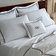 A neatly made bed with white sheets, pillowcases, and shams, all featuring gray trim. One decorative pillow in the center has a monogrammed “M” on it. The bed has a wooden headboard.