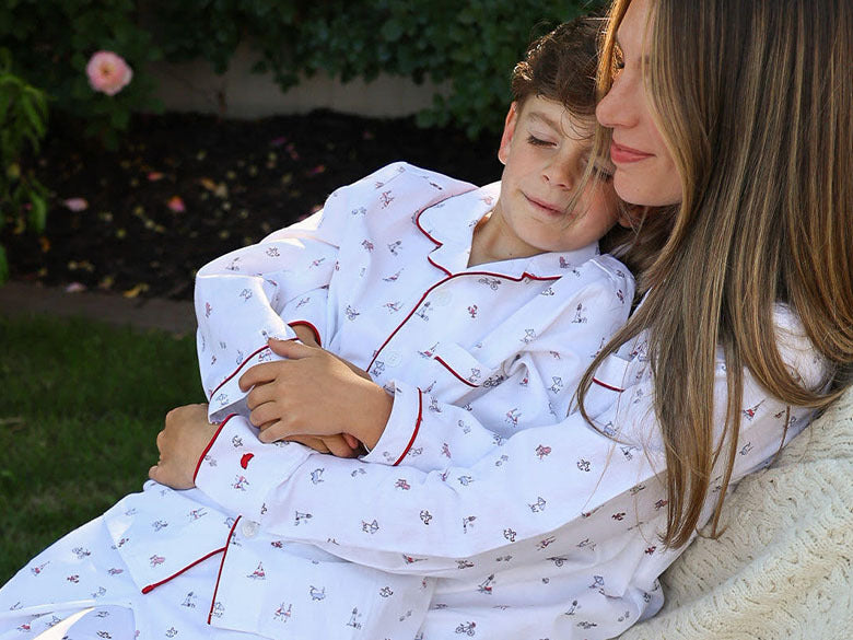 A woman and a child, both wearing matching white pajamas with red trim, sit close together outdoors. The child leans in, eyes closed, hugging the woman, who smiles gently. Greenery and flowers are in the background.