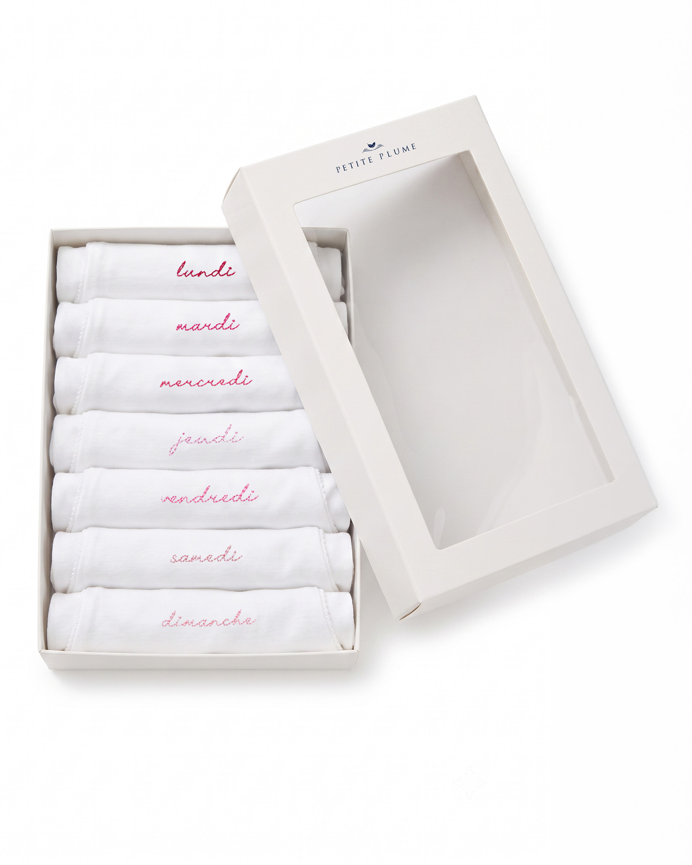 A set of seven white Baby’s Pima Long Sleeve Onesies from the “French Lessons Pink” collection, each embroidered in pink with a day of the week in French, neatly arranged in an open cream box with a clear lid.