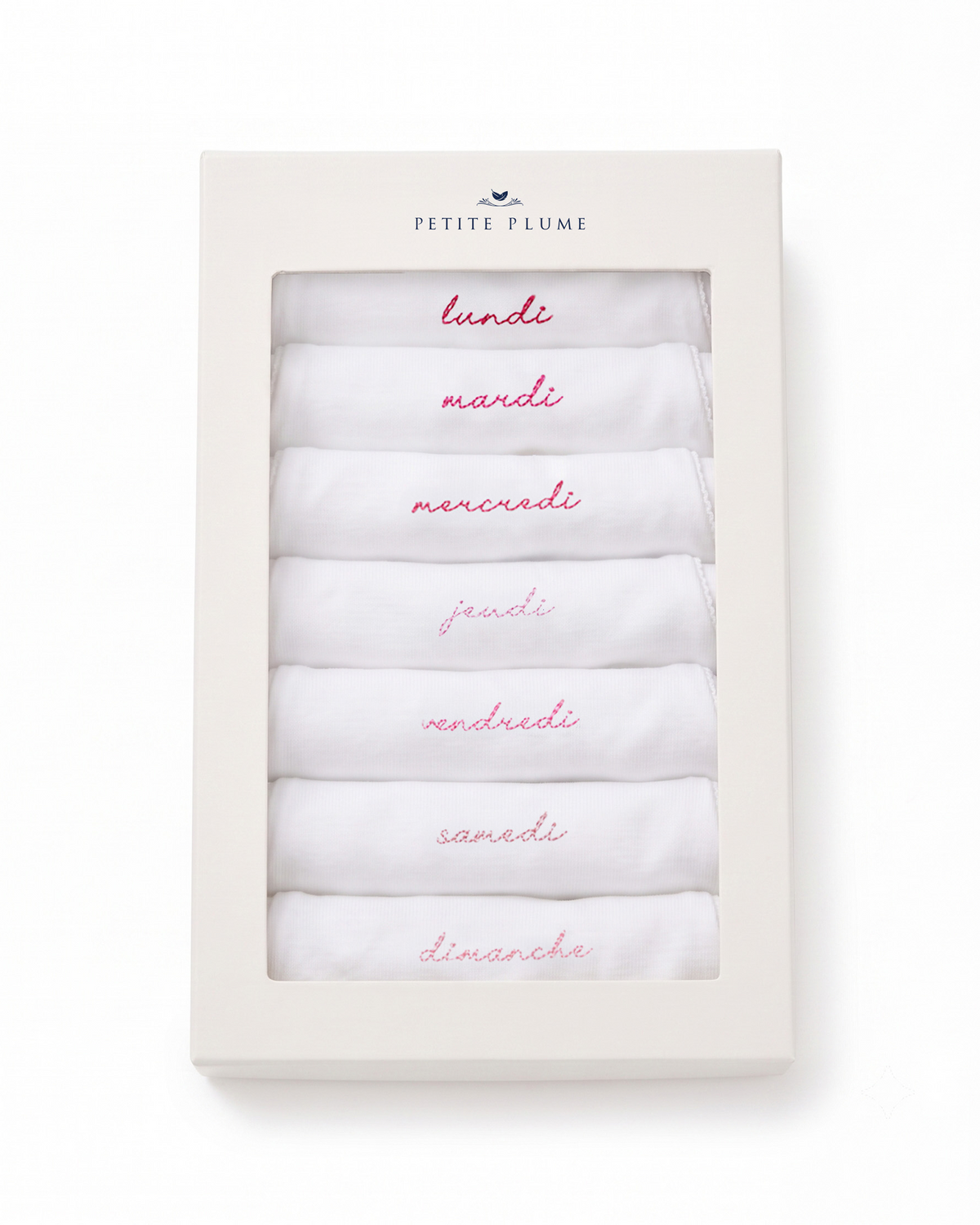 A boxed set from Petite Plume contains seven white Baby’s Pima Long Sleeve Onesies in French Lessons Pink, each embroidered with a day of the week in French and arranged in order from lundi (Monday) to dimanche (Sunday). The Petite Plume brand name appears on the box.