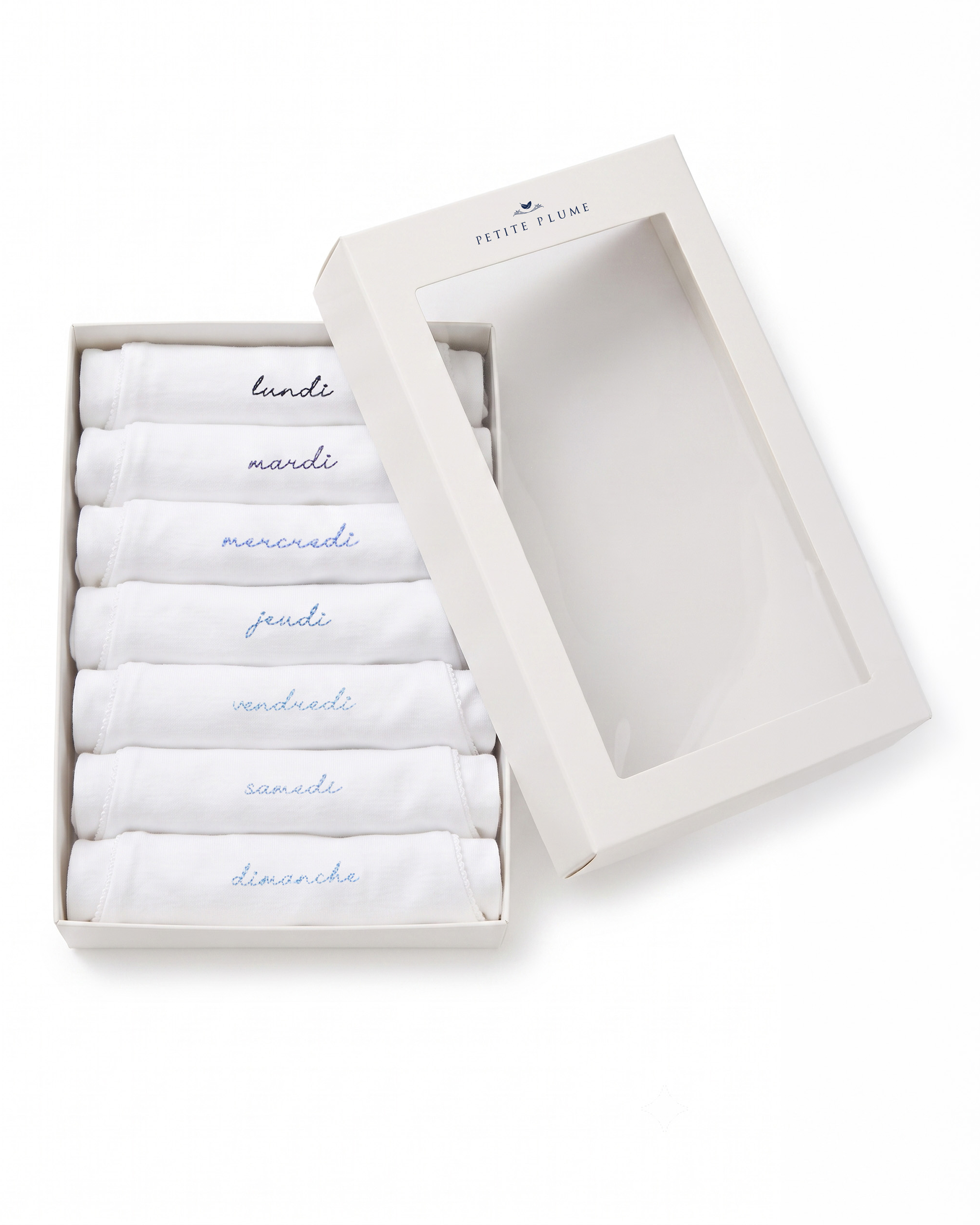 A white box with a clear lid showcases the Baby’s Pima Short Sleeve Onesie Set in French Lessons Blue, featuring seven neatly folded white onesies, each labeled with a day of the week in French: lundi, mardi, mercredi, jeudi, vendredi, samedi, and dimanche.