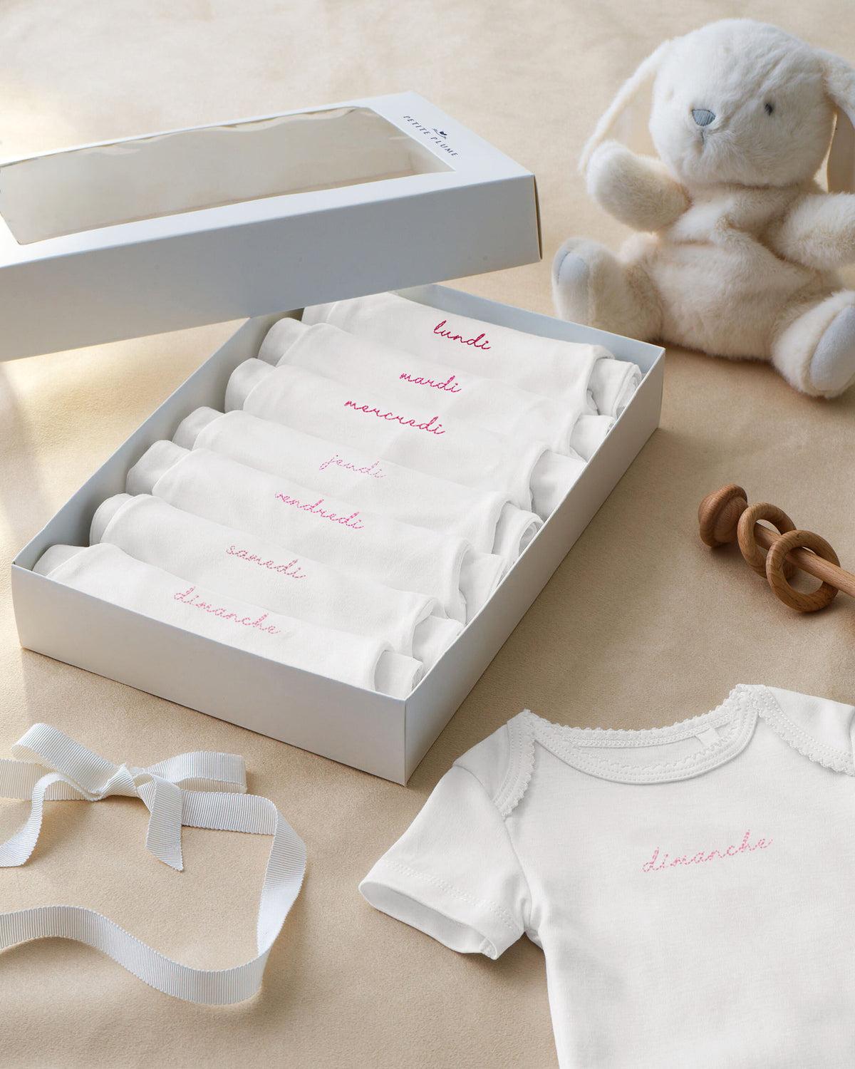 A box of seven Baby’s Pima Short Sleeve Onesie Set in French Lessons Pink—each folded and labeled with the days of the week in French—alongside a plush bunny, a wooden rattle, a ribbon, and an unfolded onesie reading “dimanche,” are arranged on a beige surface.