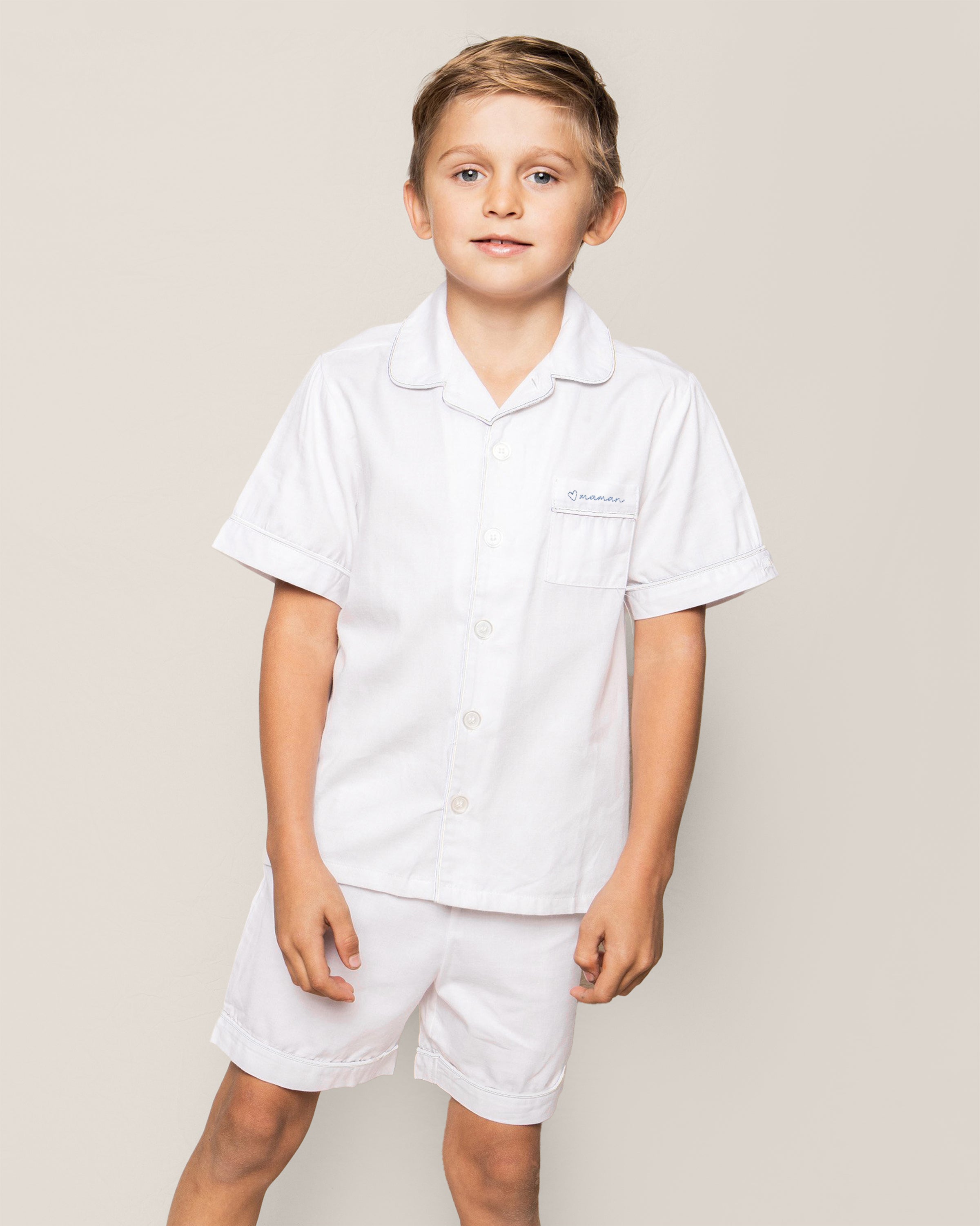 Close-up of the Kid's Twill Pajama Short Set featuring a white top with white buttons, a chest pocket embroidered with a blue heart and "maman", plus a PETITE PLUME label inside the collar.