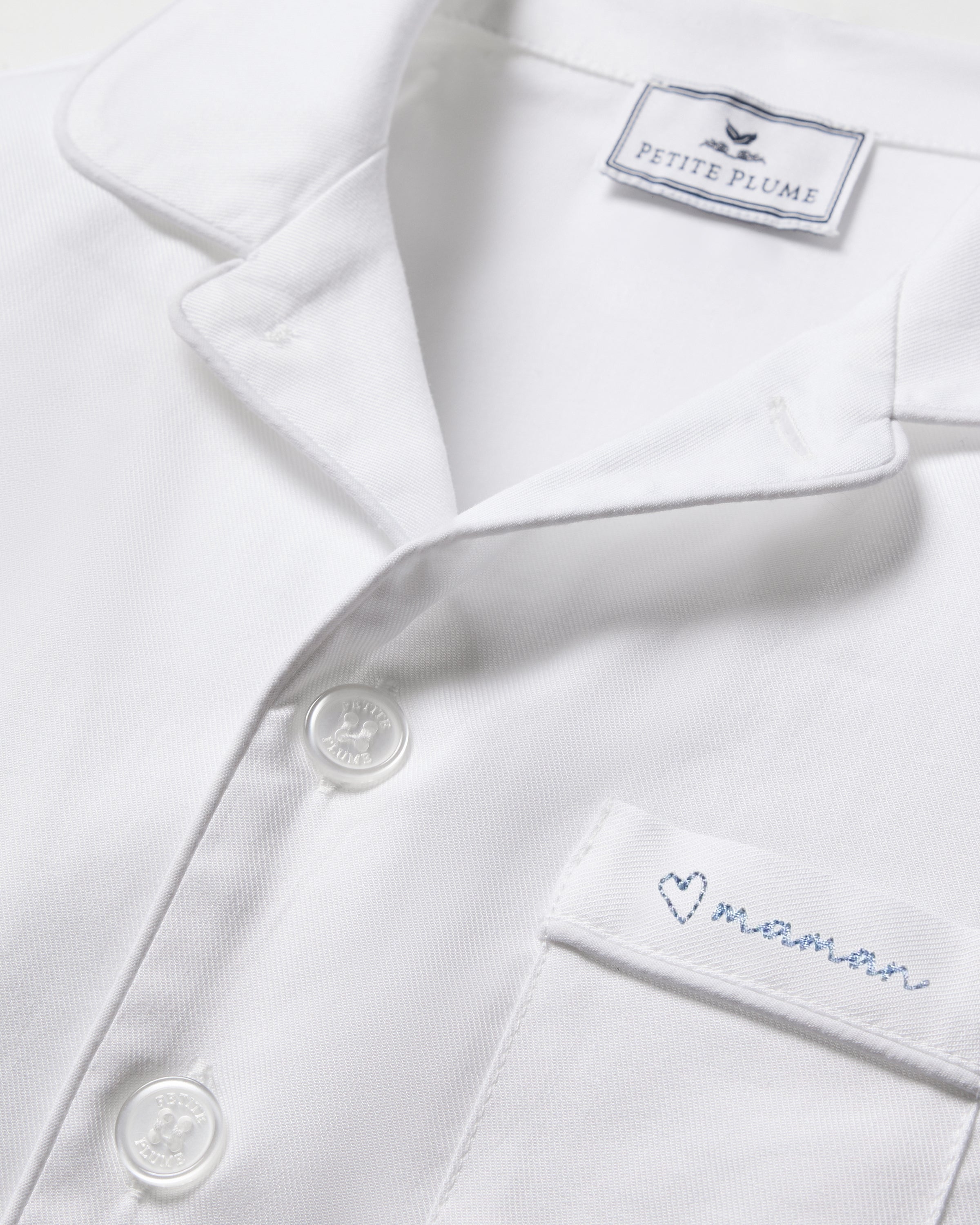 Close-up of the Kid's Twill Pajama Short Set featuring a white top with white buttons, a chest pocket embroidered with a blue heart and "maman", plus a PETITE PLUME label inside the collar.
