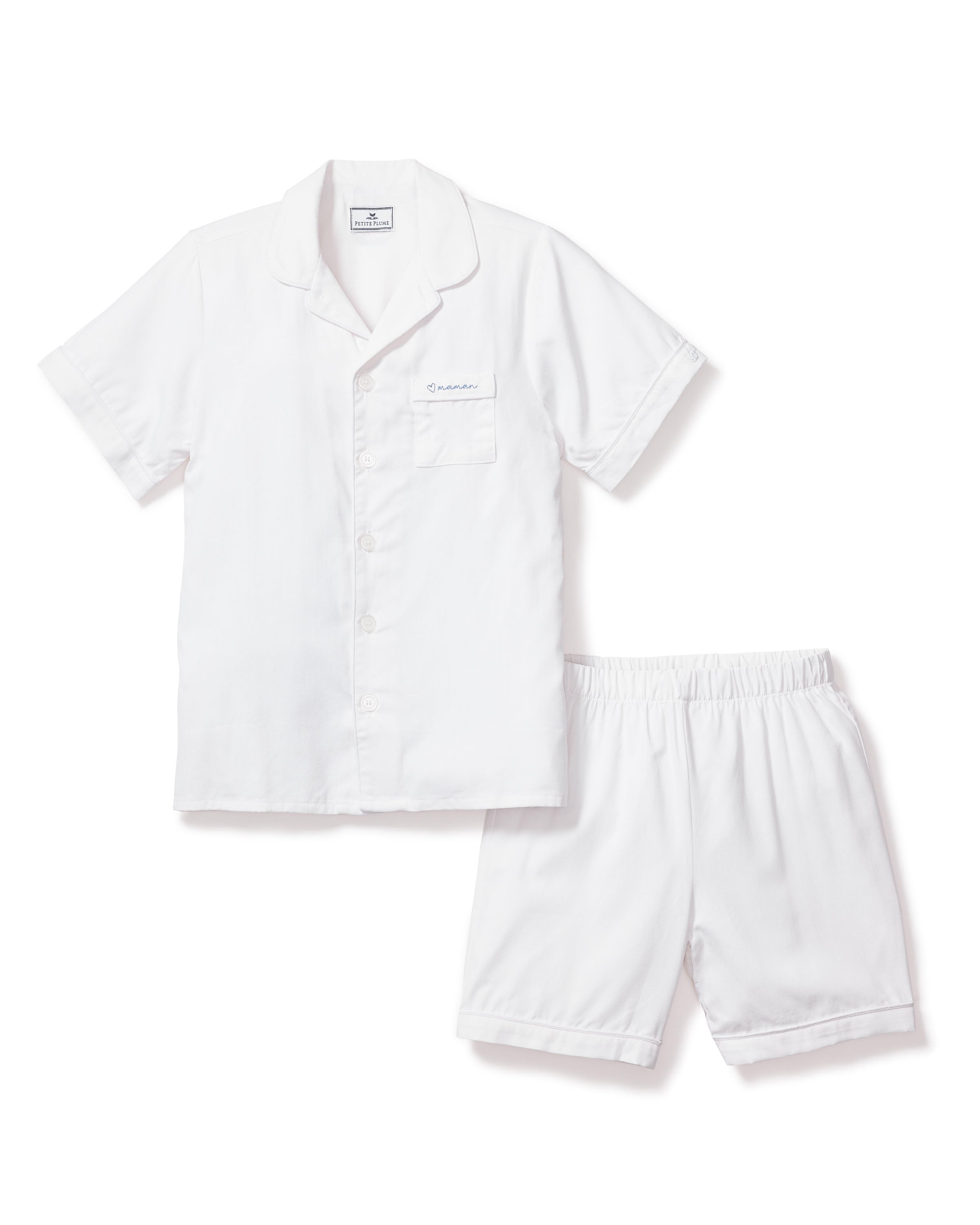 Close-up of the Kid's Twill Pajama Short Set featuring a white top with white buttons, a chest pocket embroidered with a blue heart and "maman", plus a PETITE PLUME label inside the collar.