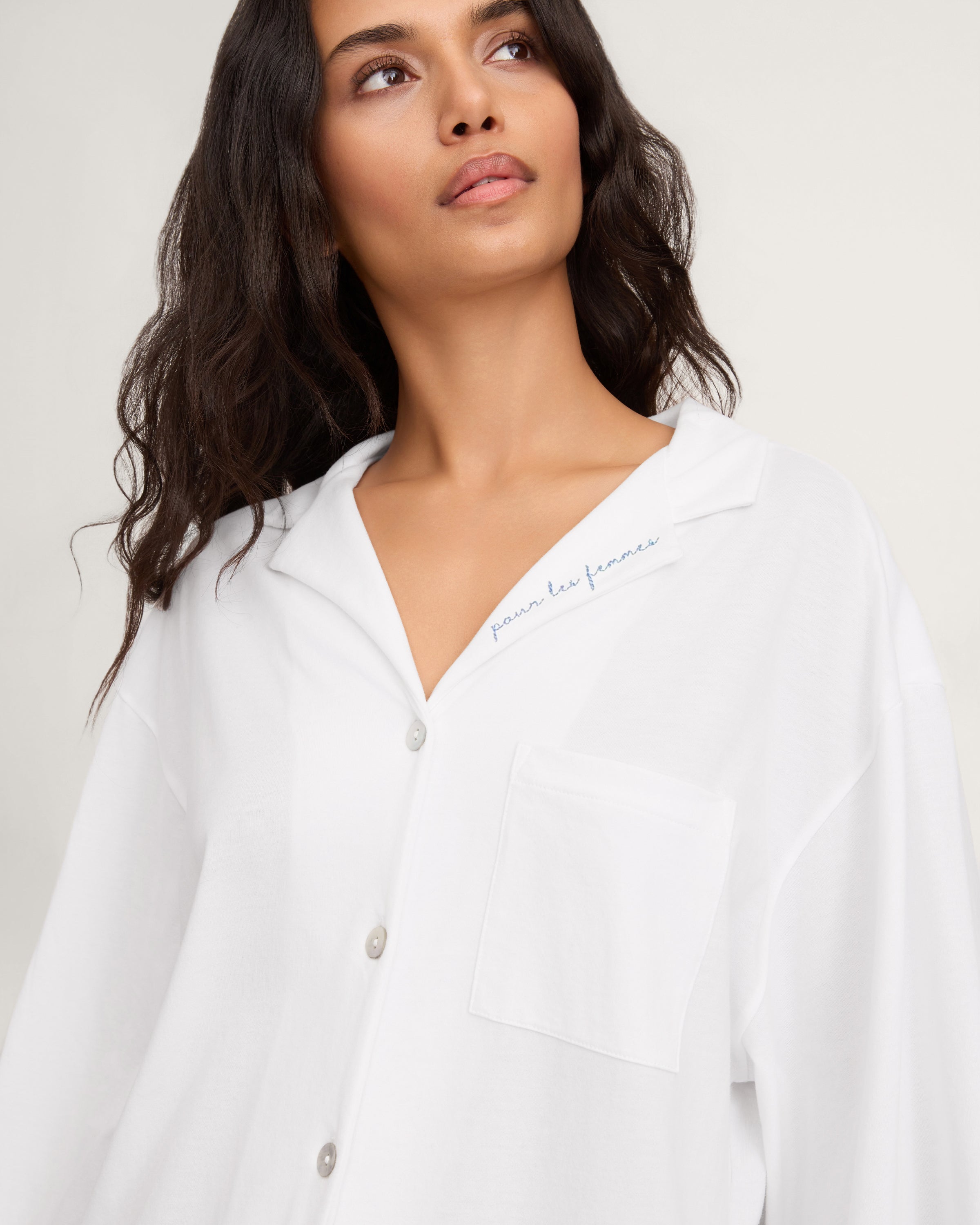 A woman with long, wavy dark hair models The Olivia Wide Leg Pima Pajama Set With "Pour Les Femmes" Embroidery, featuring a loose white button-up shirt with collar and front pocket, gazing slightly upward against a plain background.