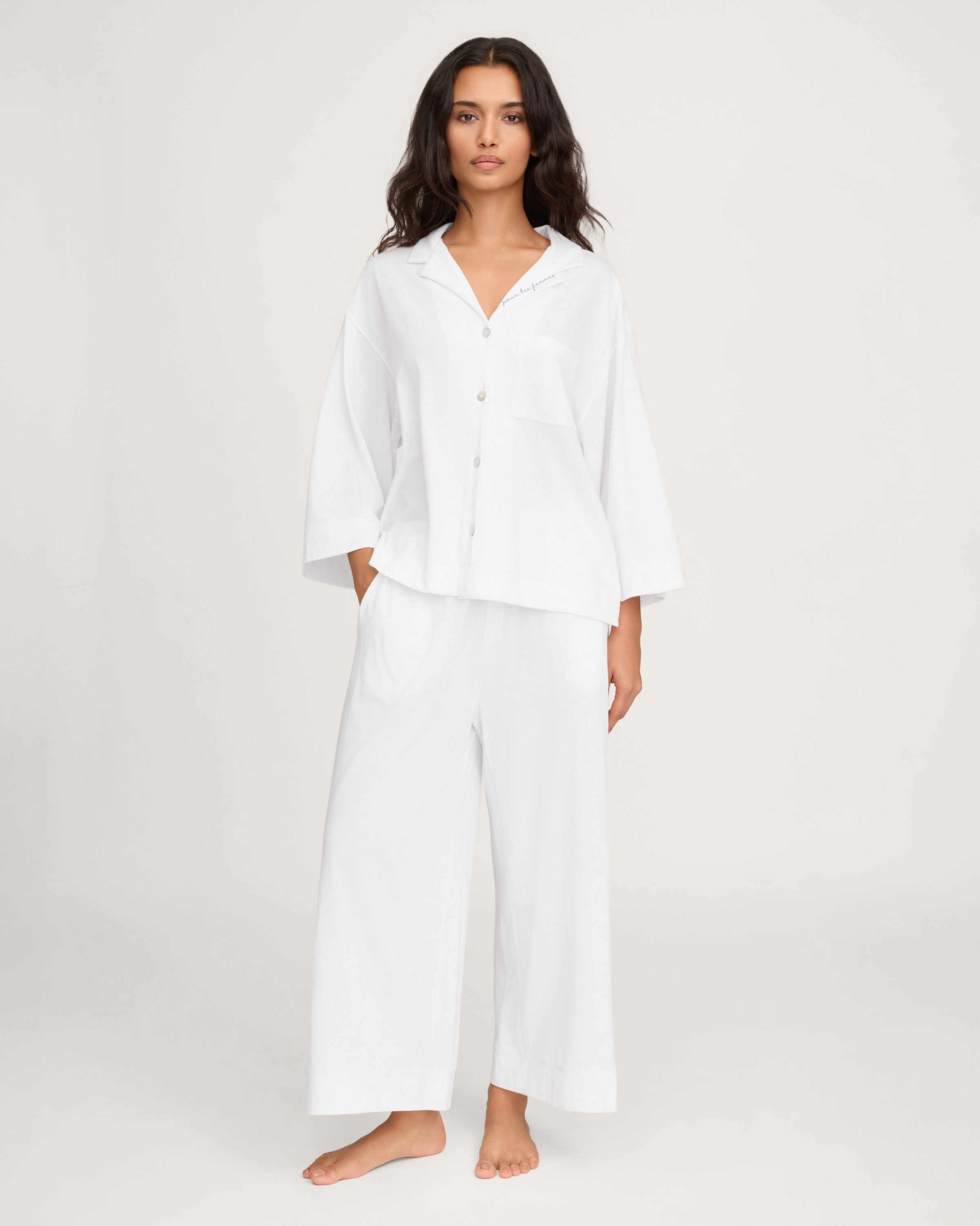 A woman with long, wavy dark hair models The Olivia Wide Leg Pima Pajama Set With "Pour Les Femmes" Embroidery, featuring a loose white button-up shirt with collar and front pocket, gazing slightly upward against a plain background.