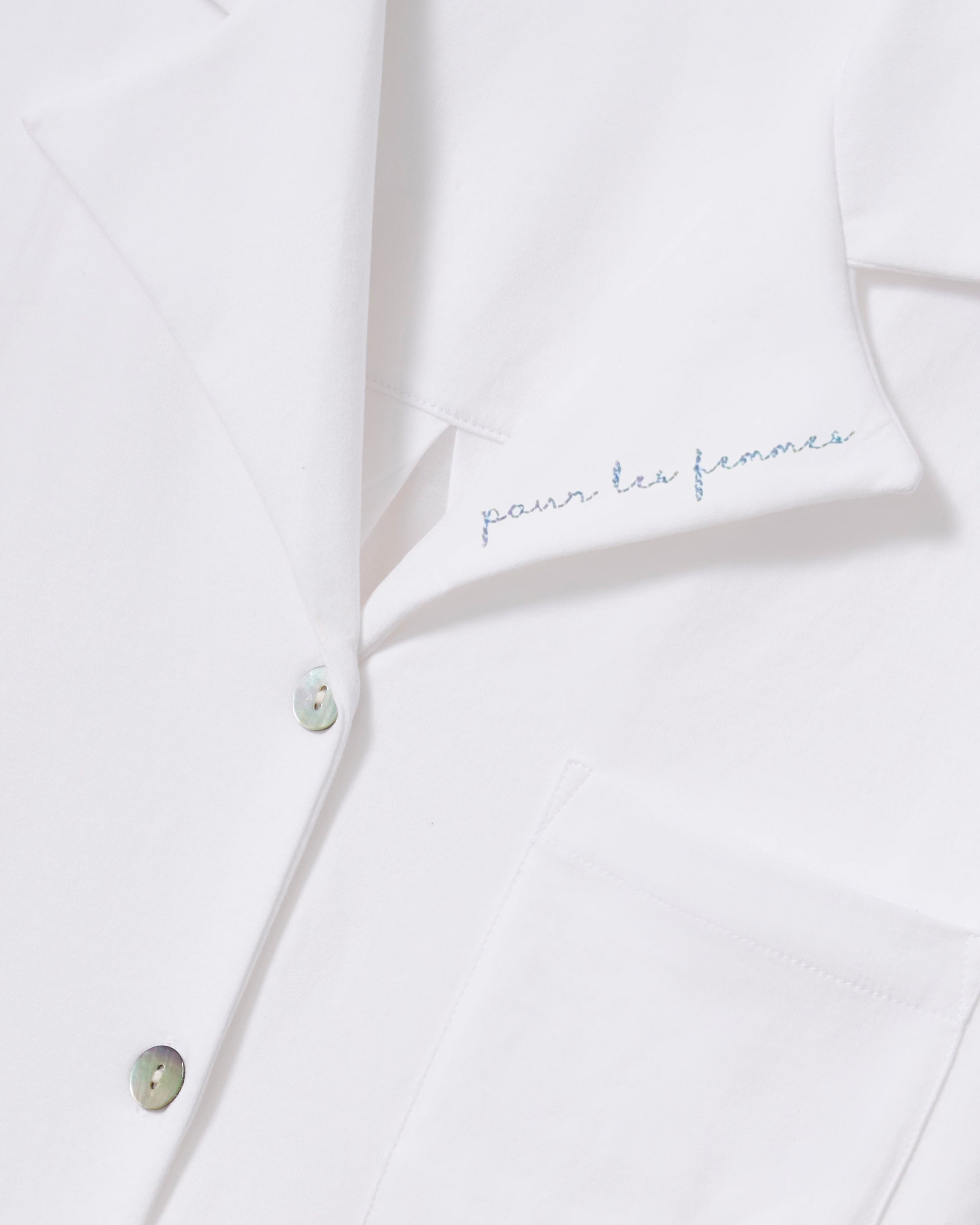 A woman with long, wavy dark hair models The Olivia Wide Leg Pima Pajama Set With "Pour Les Femmes" Embroidery, featuring a loose white button-up shirt with collar and front pocket, gazing slightly upward against a plain background.