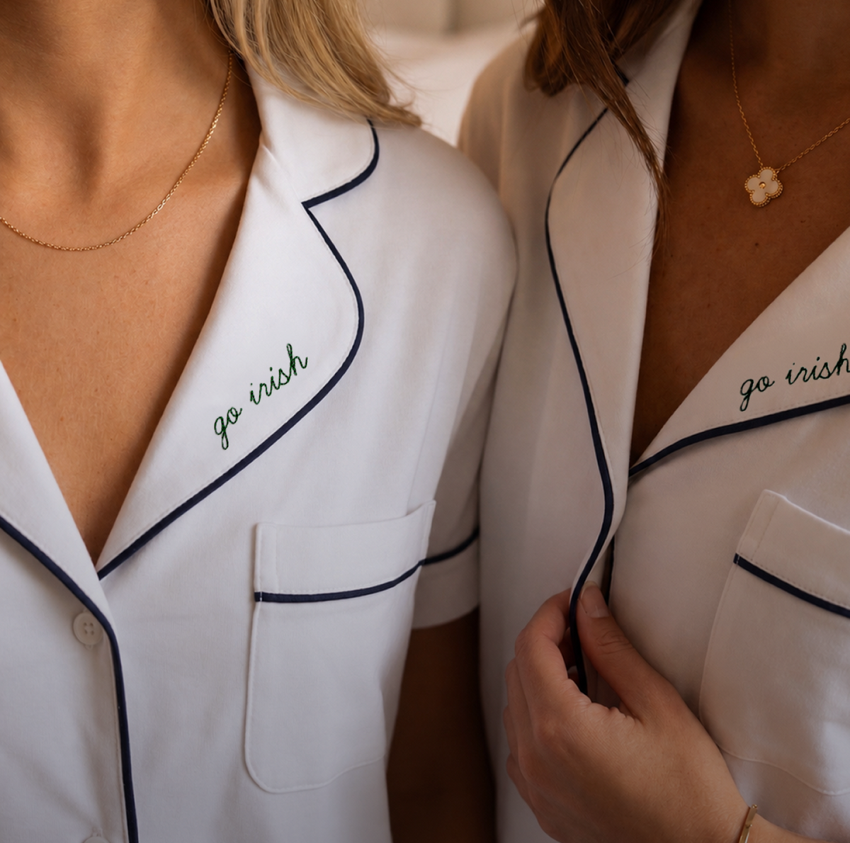 Two women wearing white pajamas with dark piping and gold necklaces. Go Irish is embroidered in green on each pajama pocket. One woman holds her collar, and another wears a flower-shaped pendant.