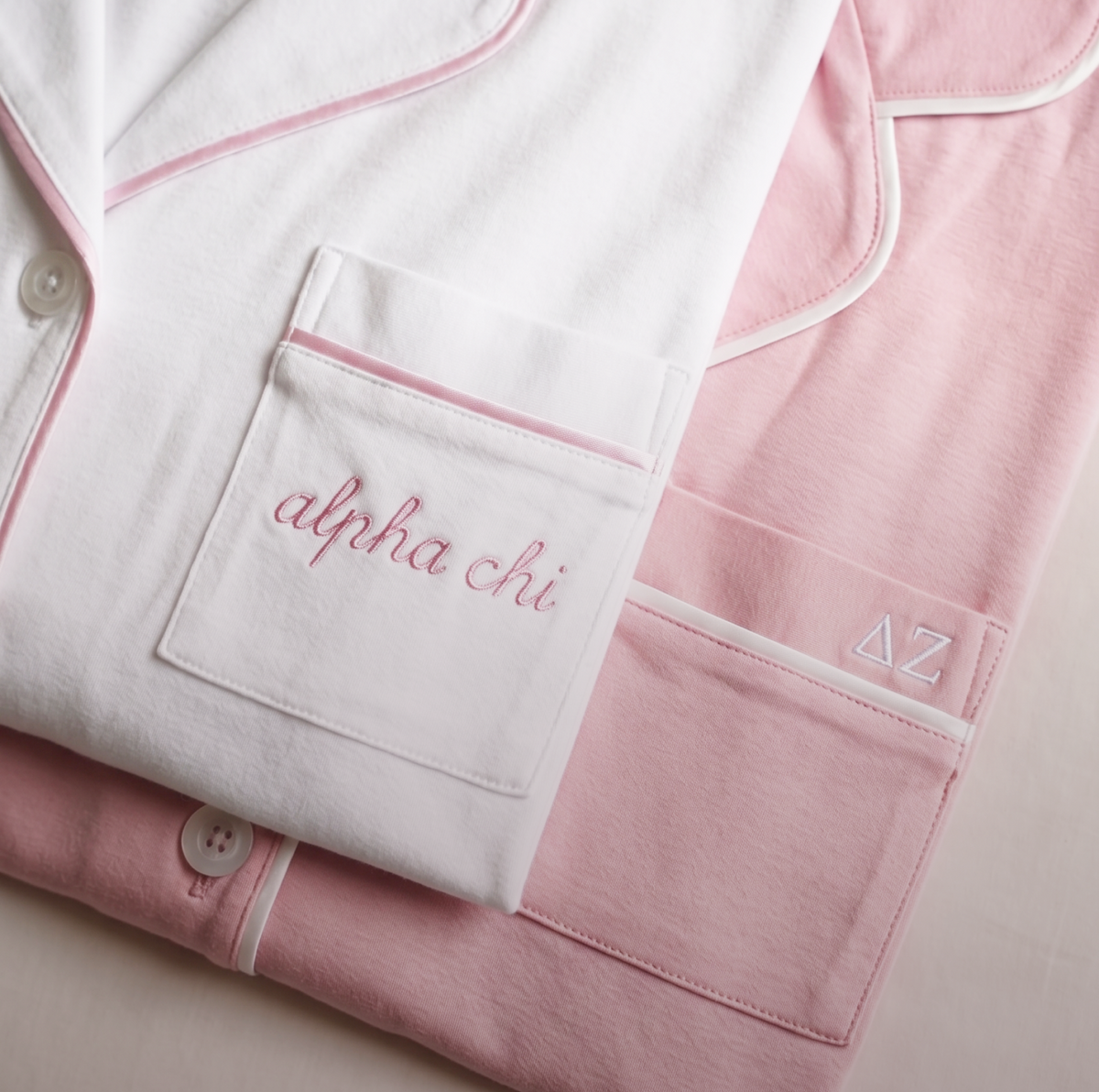 Two folded pajama tops: one white with alpha chi embroidered on the pocket, and one pink with the Greek letters ΔΖ on the pocket, both with pink piping along the edges.