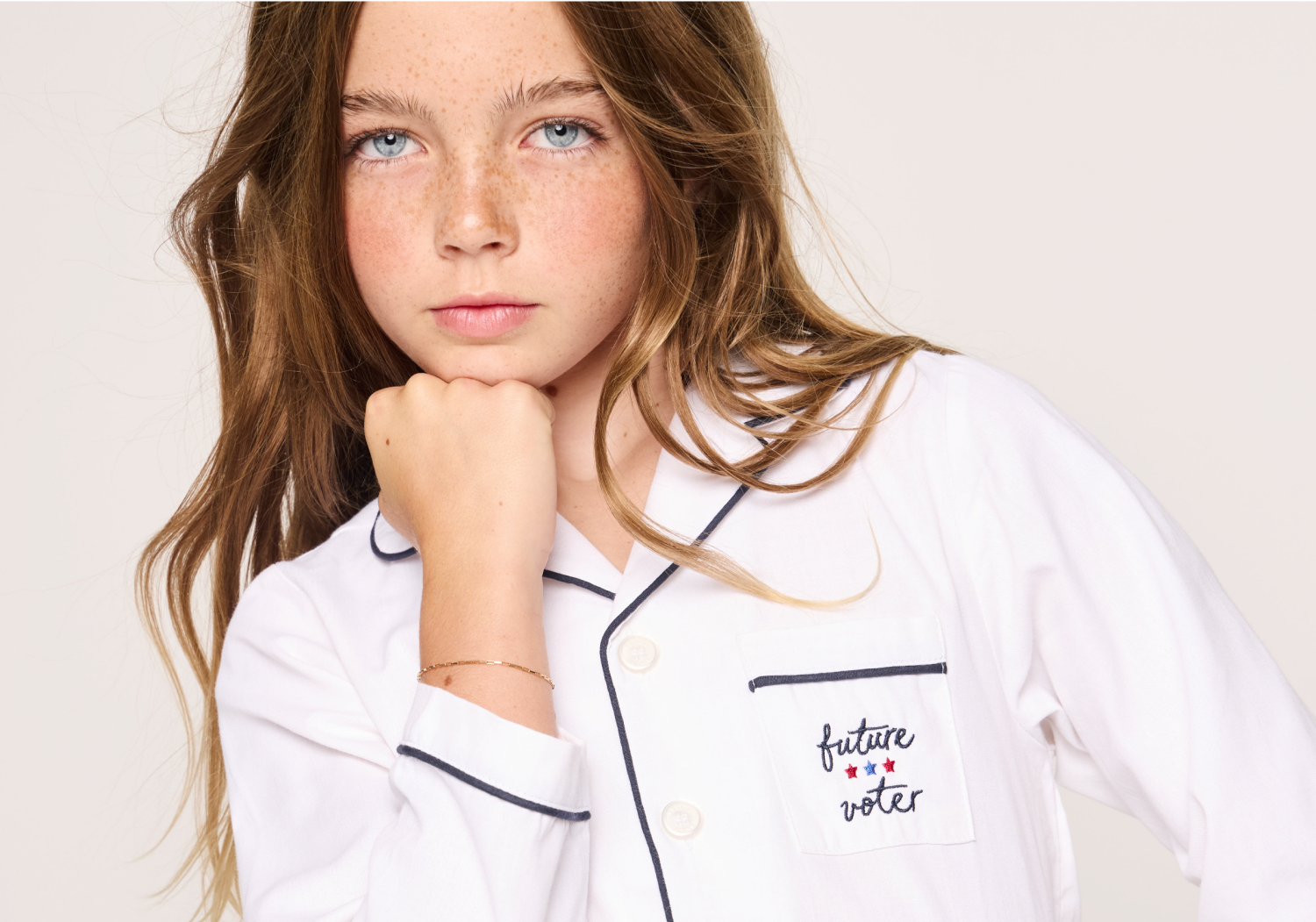 A young girl with long brown hair and blue eyes rests her chin on her hand. She wears white pajamas with black piping and a pocket embroidered with the words “future voter” and two red stars.