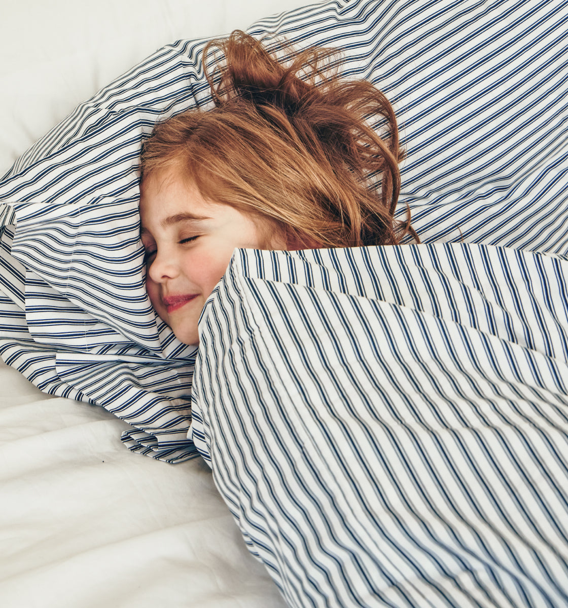 A young child with brown hair lies in bed, smiling peacefully while sleeping, covered with a white and blue striped blanket and pillow.