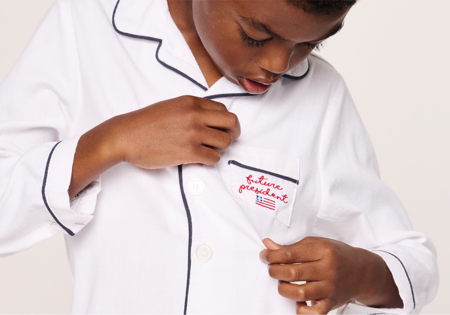 A child in white pajamas with black piping looks down and points at a chest pocket embroidered with an American flag and the words “future president” in red.