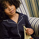 A young child with curly hair, wearing dark pajamas with white trim, sits on a striped chair holding a fabric object, looking slightly upward with a gentle expression.
