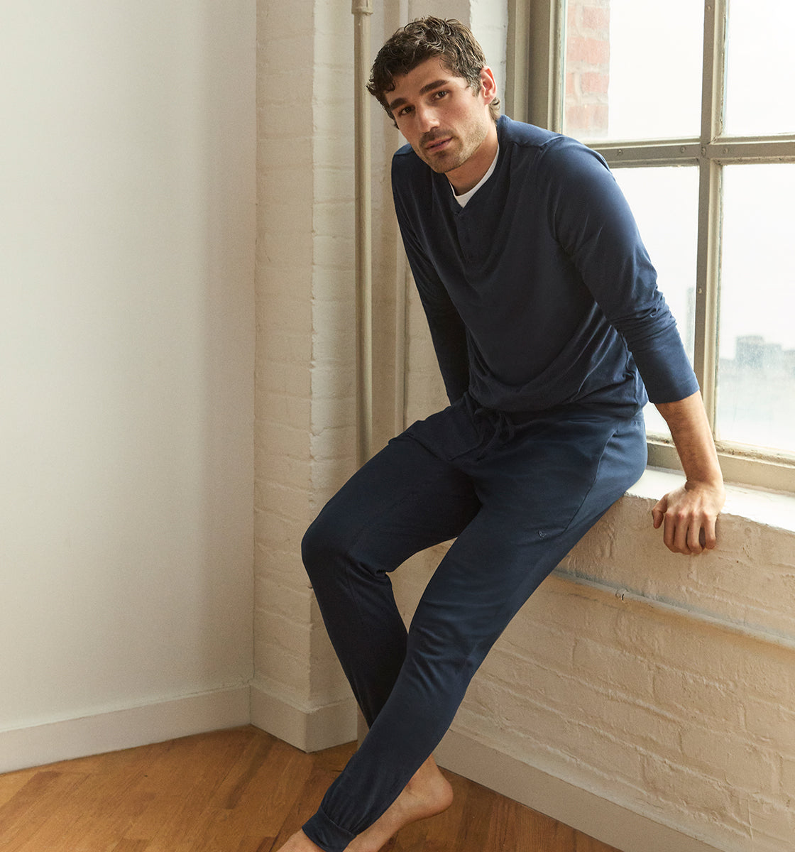 A man wearing a navy blue long-sleeve shirt and matching pants sits barefoot on a windowsill in a bright room with white walls and hardwood floors.