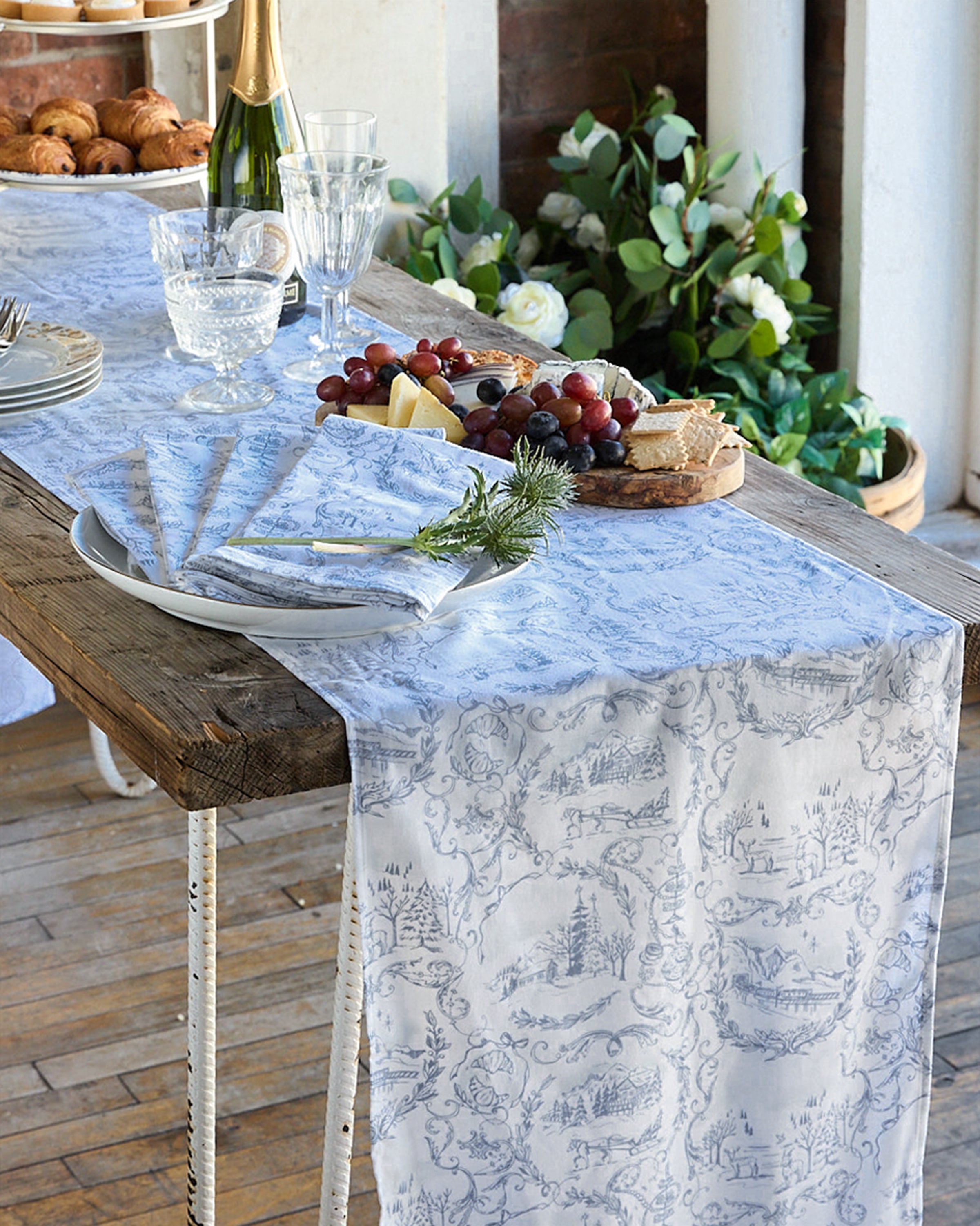 A rustic wooden table is elegantly set with 100% cotton Maman x Petite Plume Alpine Toile Runner, plates, cutlery, and glasses. A platter of cheese, grapes, bread, a bottle of champagne, and a vase of flowers complete the festive tablescape.