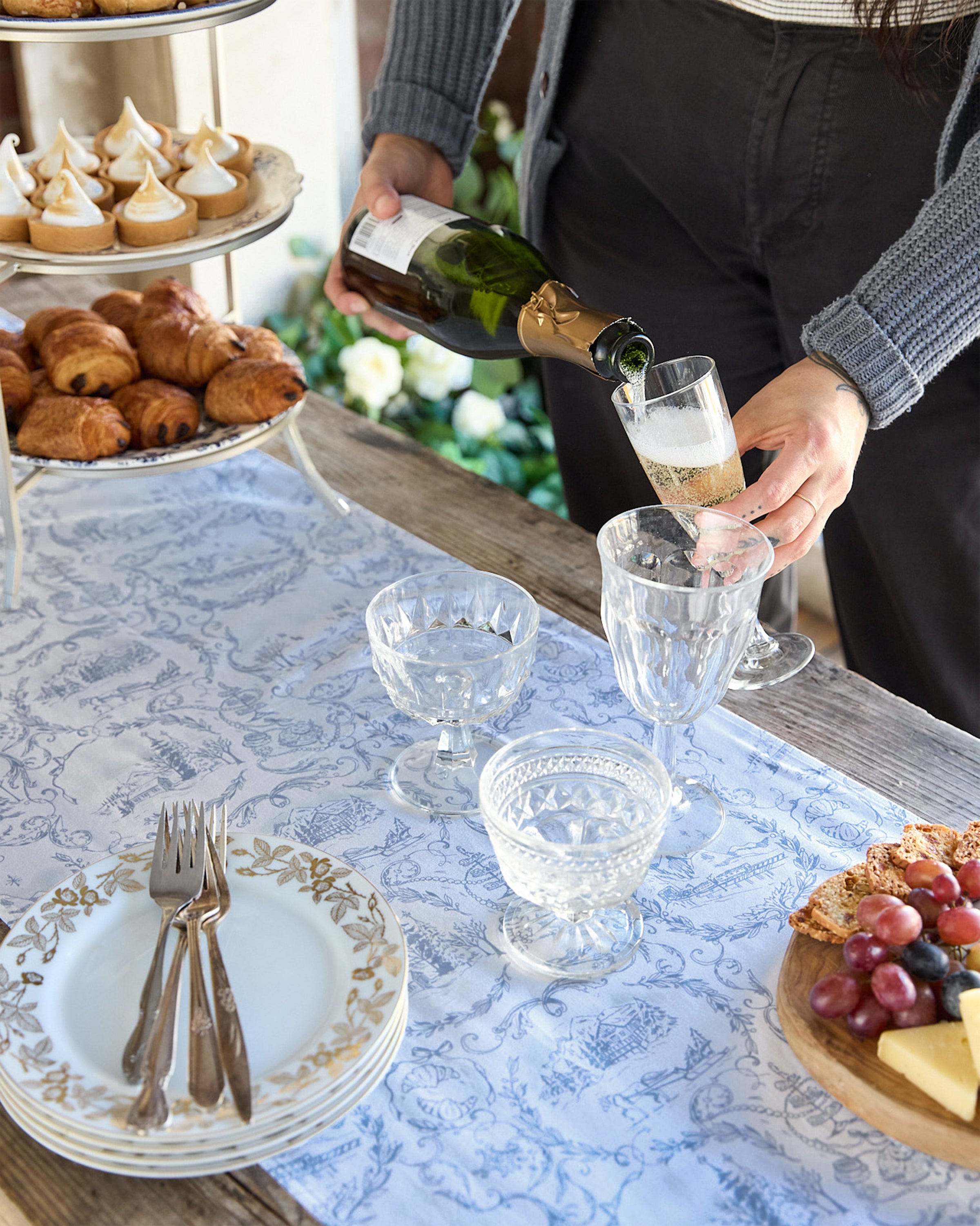Someone pours champagne into a tall glass beside a festive setting featuring desserts on a tiered stand, plates with forks, and a platter with cheese, grapes, and bread, all elegantly displayed atop the Maman x Petite Plume Alpine Toile Runner by Petite Plume. The scene exudes celebration or relaxed gathering vibes.