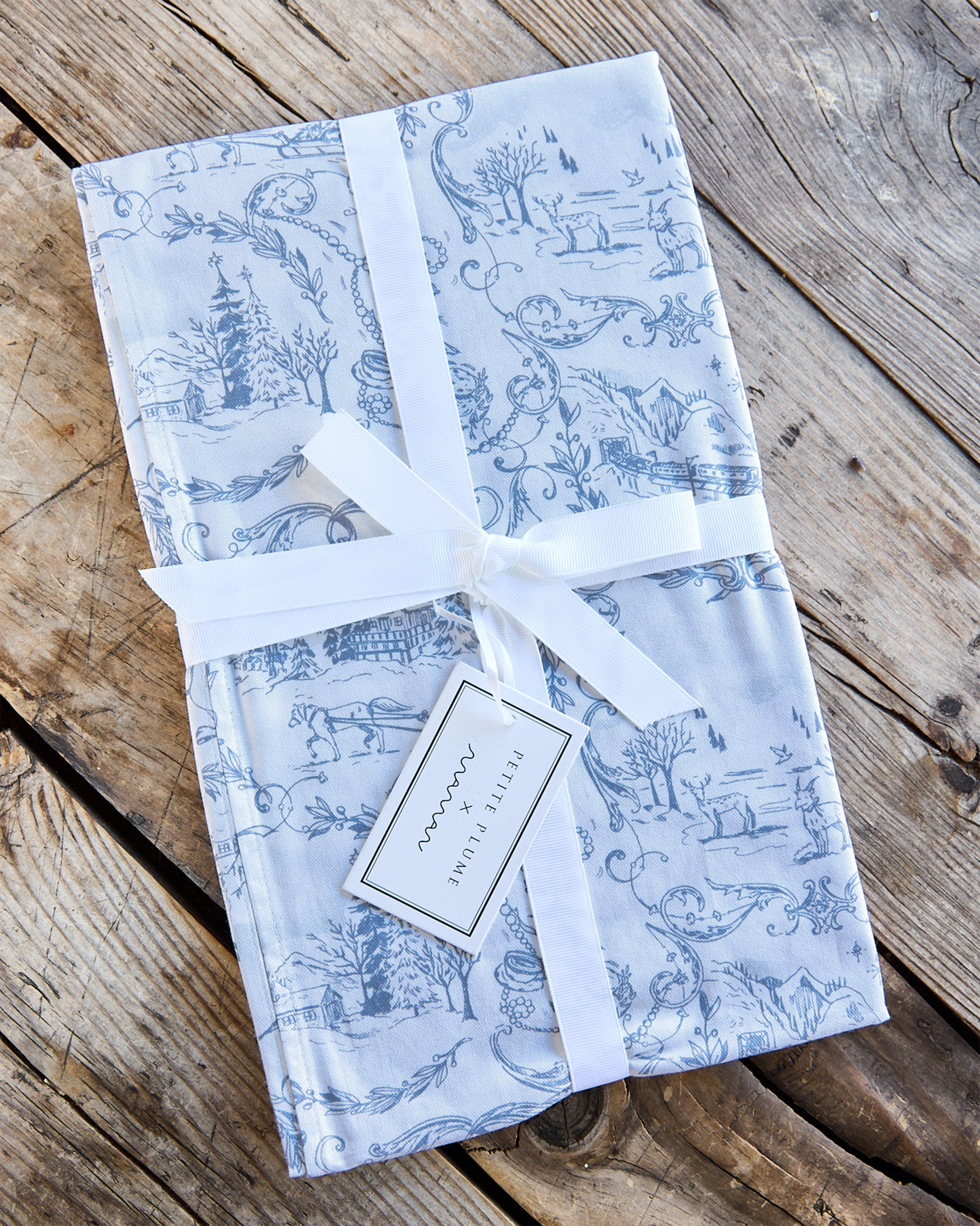 Resting on a rustic wooden surface, the Maman x Petite Plume Alpine Toile Runner features a slate blue toile design with pastoral scenes. Elegantly tied with a white ribbon and small tag, it is perfect for enhancing your festive tablescape.