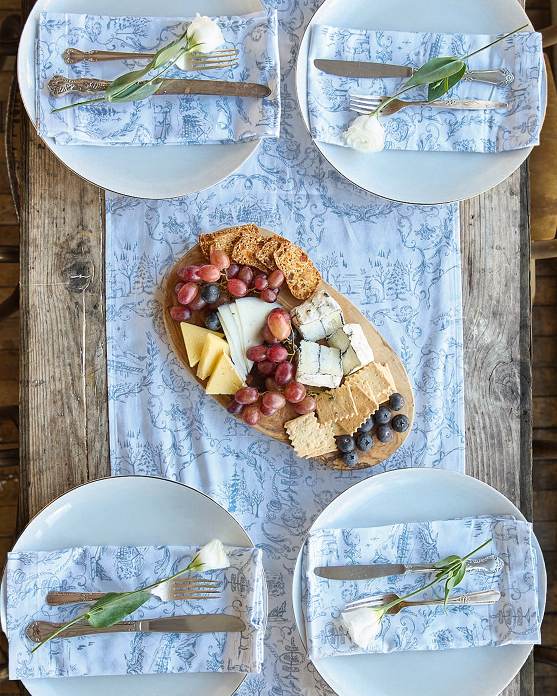 An overhead shot reveals a wooden table adorned with four plates and pure cotton linens. Each plate holds a white rose on the napkin. Centered on the tabletop is a cheese board featuring grapes, cheeses, crackers, and blueberries atop the Maman x Petite Plume Alpine Toile Runner by Petite Plume.