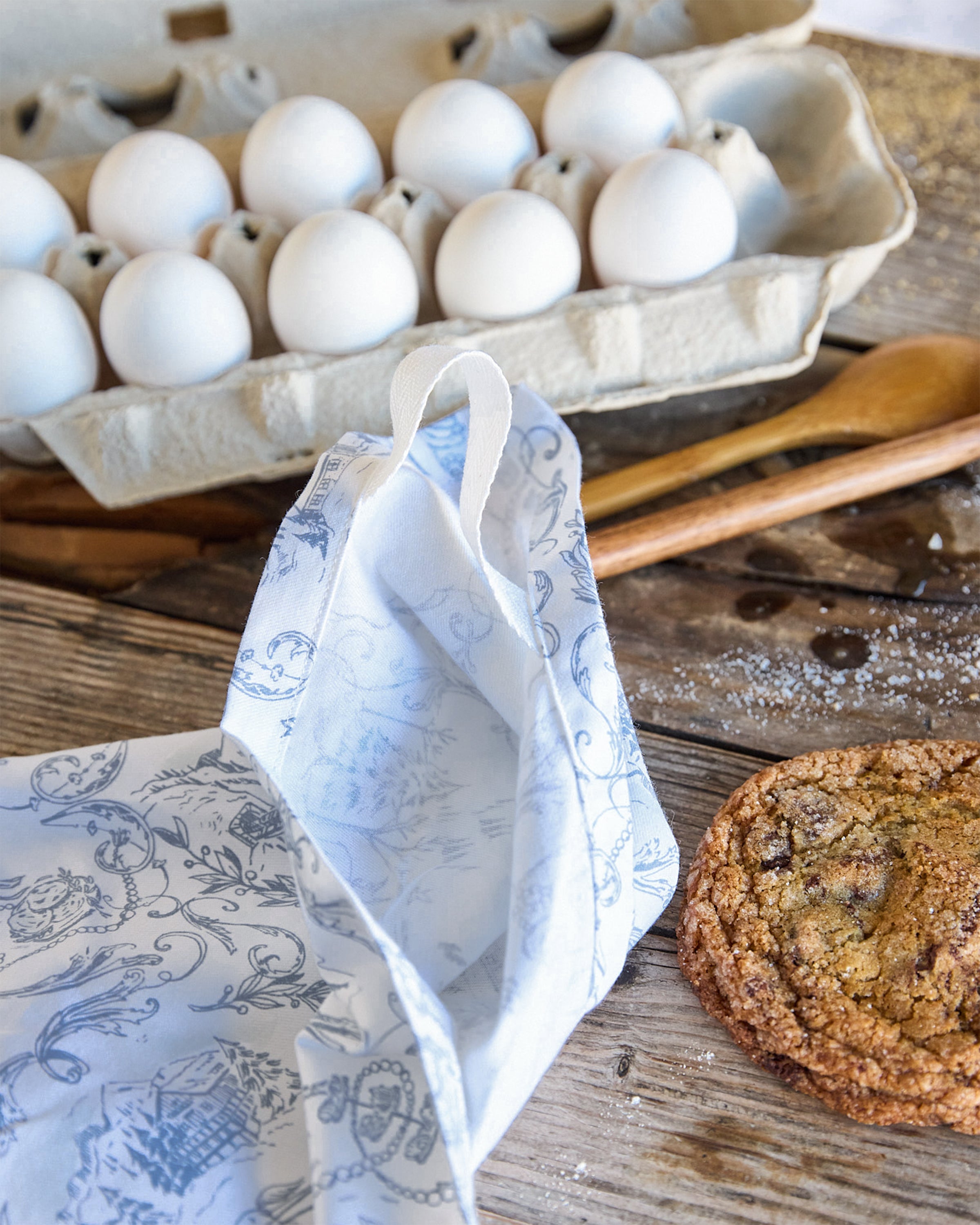 On a wooden surface, a carton of white eggs is beside a chocolate chip cookie and wooden spoon. A piece of patterned parchment paper and Maman x Petite Plume Alpine Toile Tea Towels by Petite Plume are arranged to create a cozy baking scene.