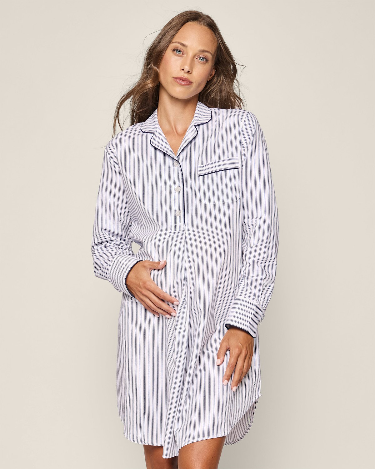 A person with long hair wears the Womens Twill Maternity Nightshirt in Navy French Ticking by Petite Plume. Against a neutral background, they look at the camera with a relaxed expression, one hand on their hip and the other on their stomach.