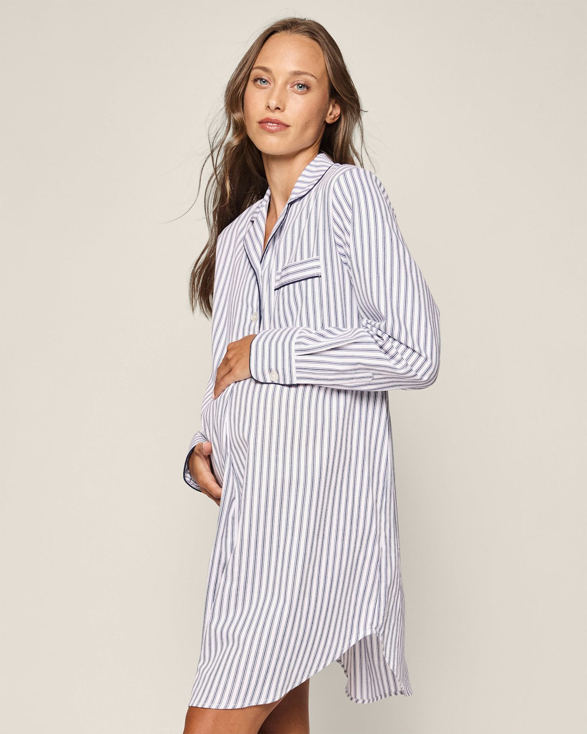 A pregnant woman with long brown hair, wearing a Womens Twill Maternity Nightshirt in Navy French Ticking by Petite Plume, gently gazes at the camera while her left hand rests on her belly against a light beige background.