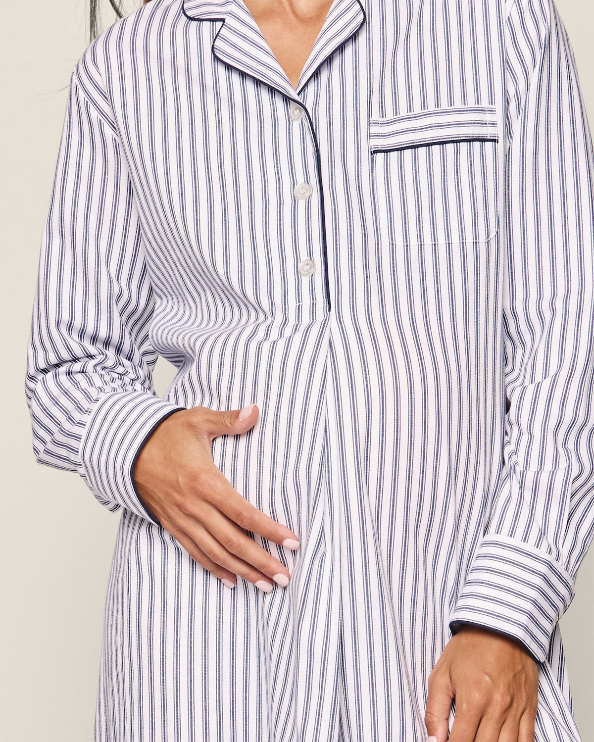 A person wearing Petite Plumes Womens Twill Maternity Nightshirt in Navy French Ticking, crafted from premium cotton with yarn-dyed dark stripes, gently touches their stomach. The nightshirt features a small chest pocket and button-up front against a light, plain background for a luxurious feel.